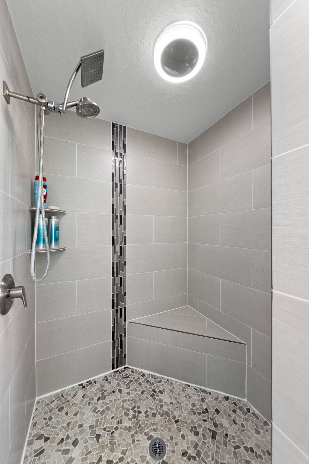 Walk-in shower featuring gray tile walls with a mosaic accent strip, a built-in corner bench, a pebble-finish floor, and a dual shower head system