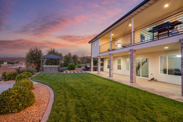 Expansive rear exterior featuring a manicured lawn, an upper-level balcony with horizontal metal railings, and a ground-level covered patio