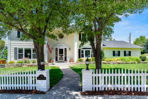 Two-story residence featuring a classic picket fence, a manicured lawn, mature trees, and a stone-lined garden
