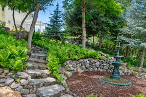 Landscaped garden featuring stone steps, mature trees, lush ferns, a decorative fountain, and a white pergola in the background