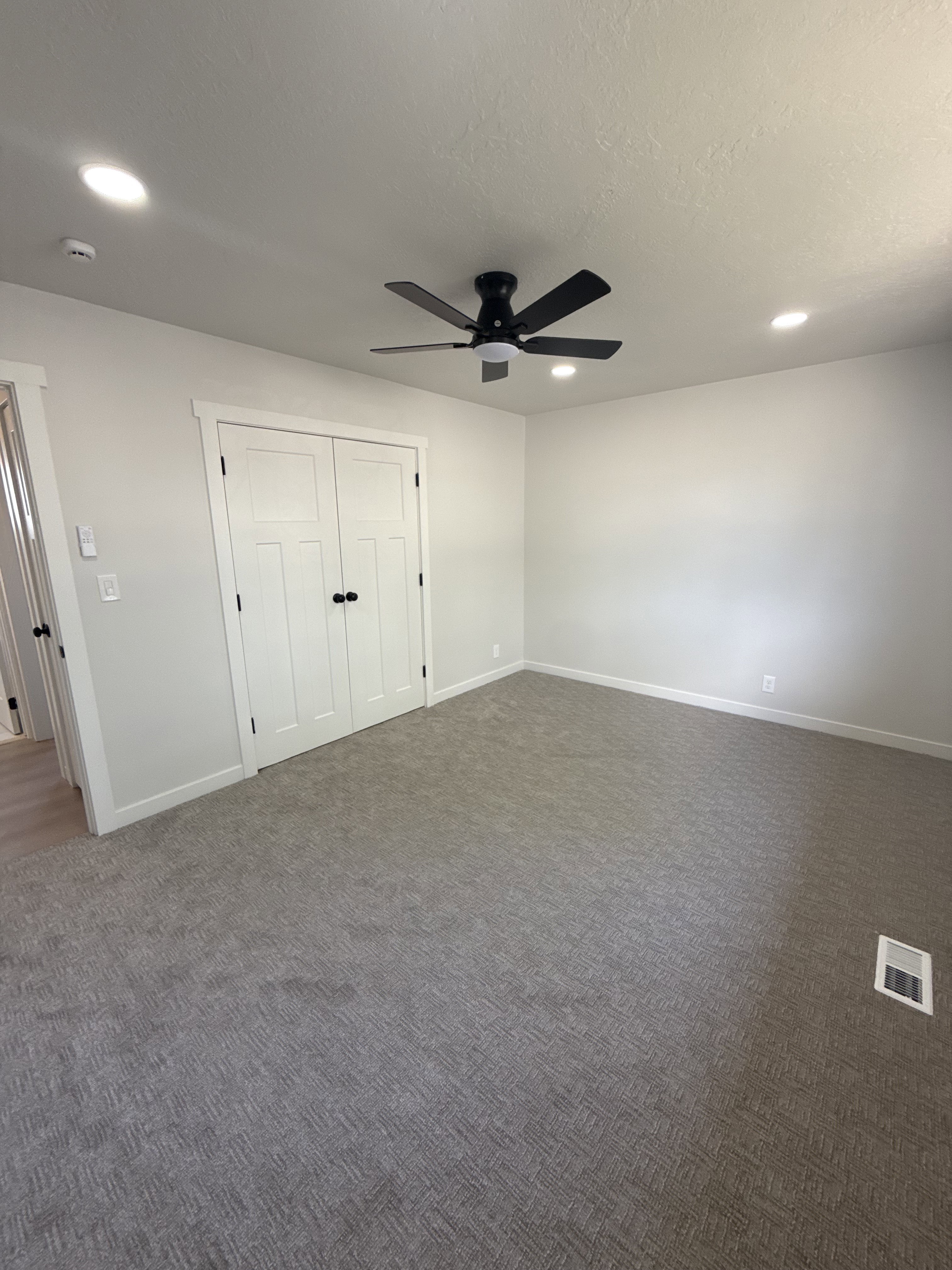 Spacious room featuring neutral carpeting, a dual-door closet with black hardware, and a black ceiling fan with integrated lighting