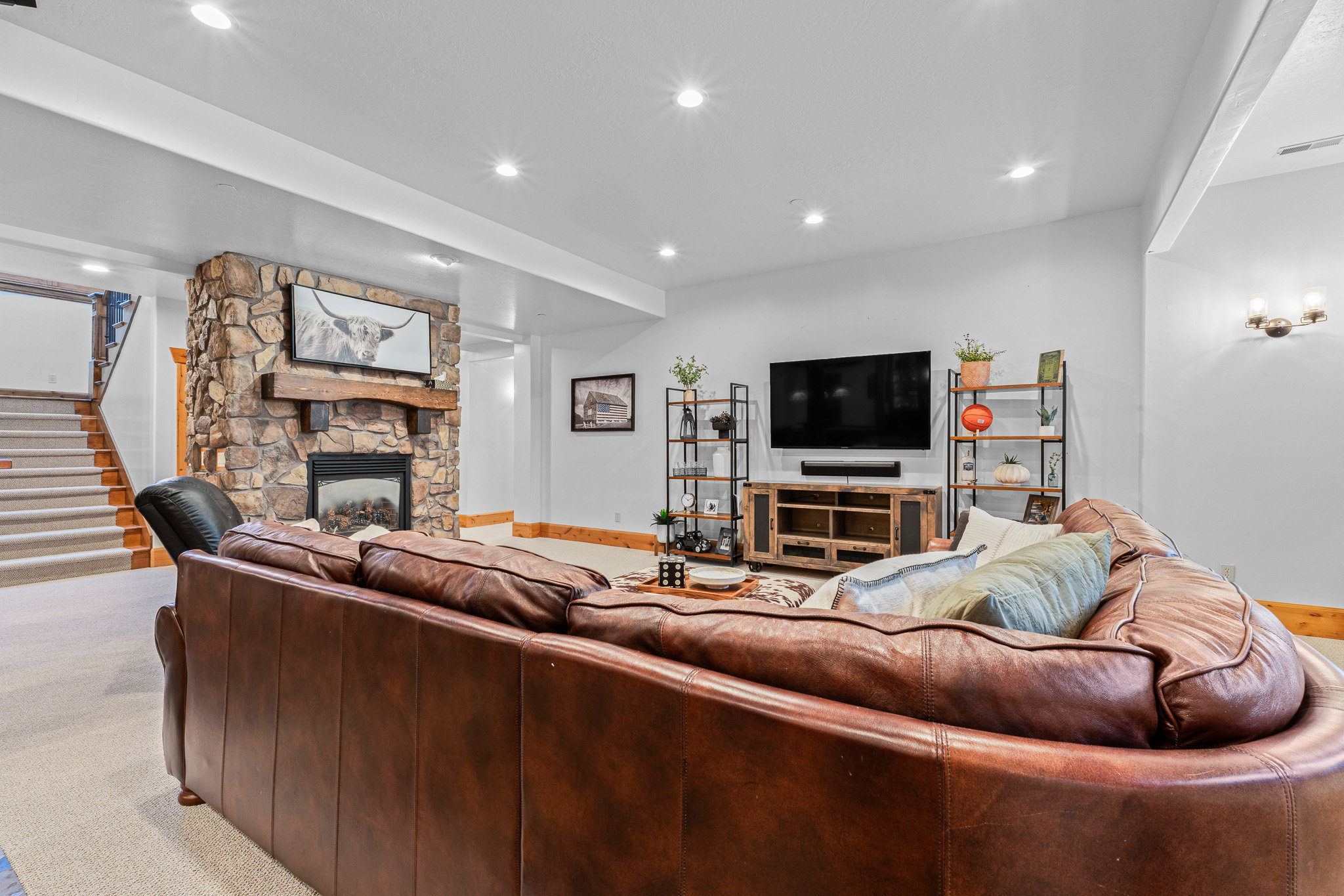 Spacious living area featuring a floor-to-ceiling stone fireplace with a wood mantel, recessed lighting, wall sconces, and a carpeted staircase