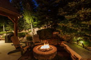 Stone fire pit with a natural stone cap, flagstone patio, covered patio structure with exposed wood beams, mature landscaping with accent lighting, and natural rock retaining walls