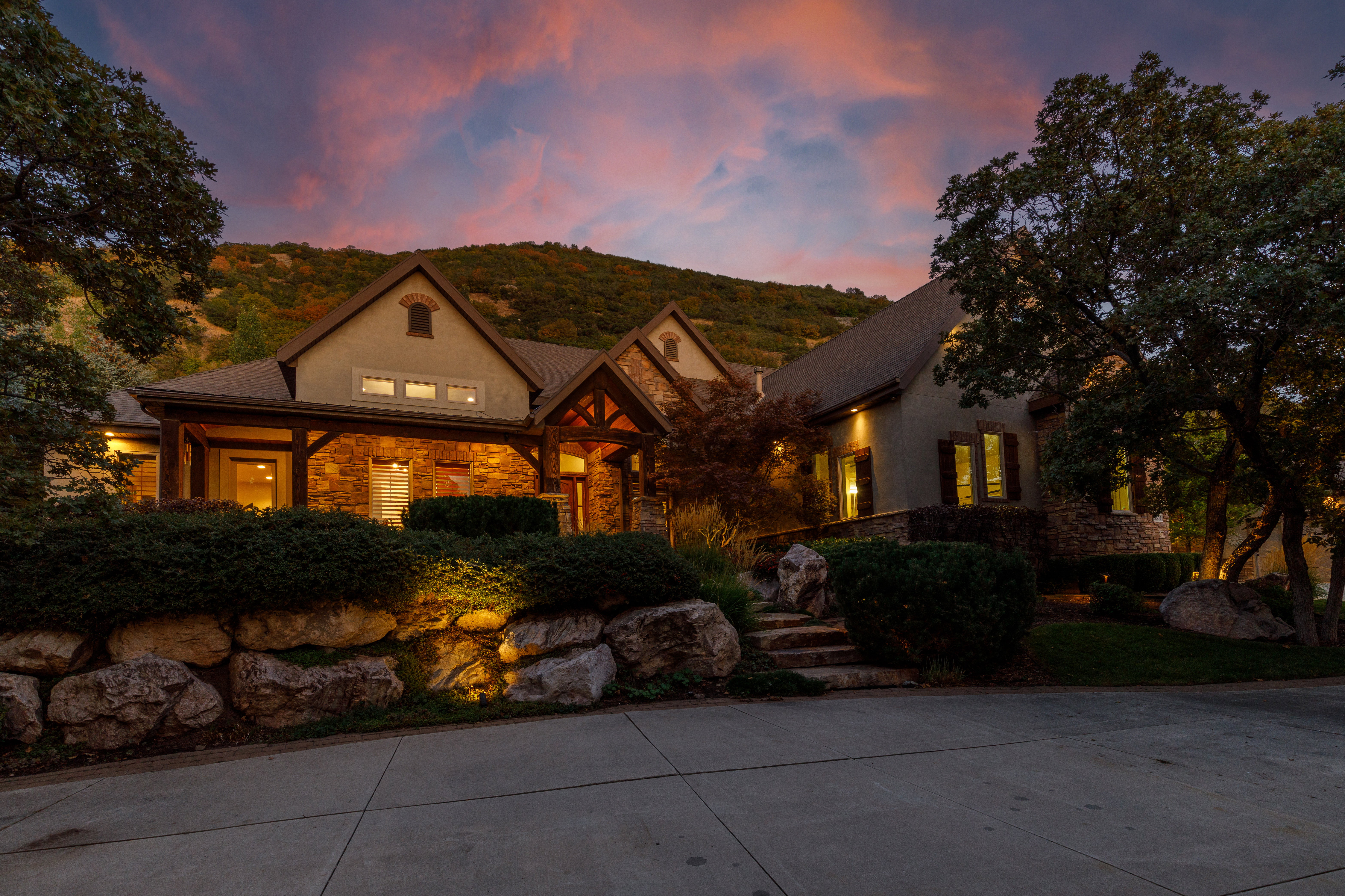 Stone and stucco exterior with exposed timber architectural accents, a multi-gabled roofline, and established landscaping featuring mature trees and rock retaining walls