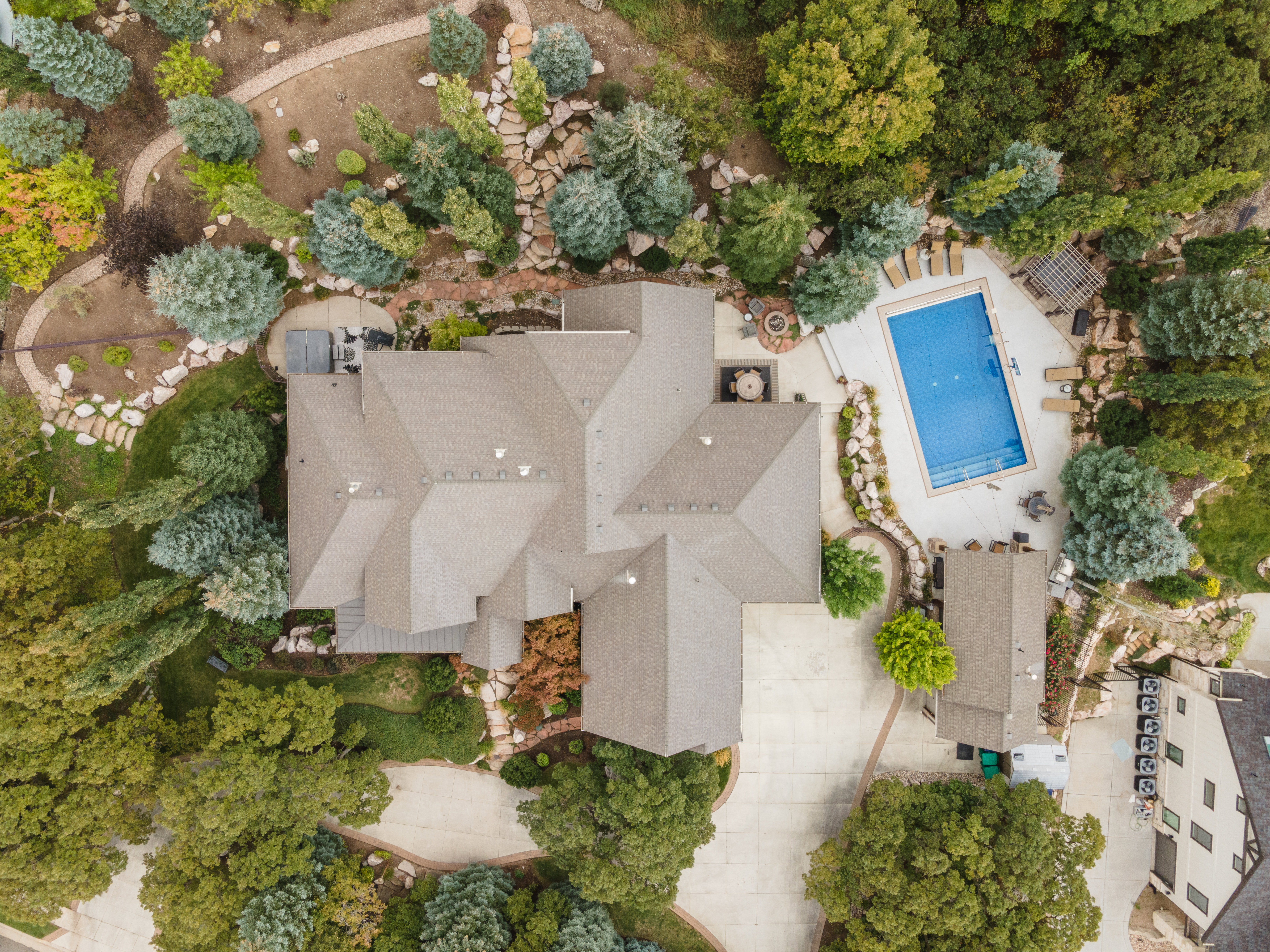 Sprawling property featuring a multi-gabled roofline, a rectangular swimming pool with surrounding patio, extensive landscaping with mature trees and stone pathways, a paved driveway, and an additional detached structure