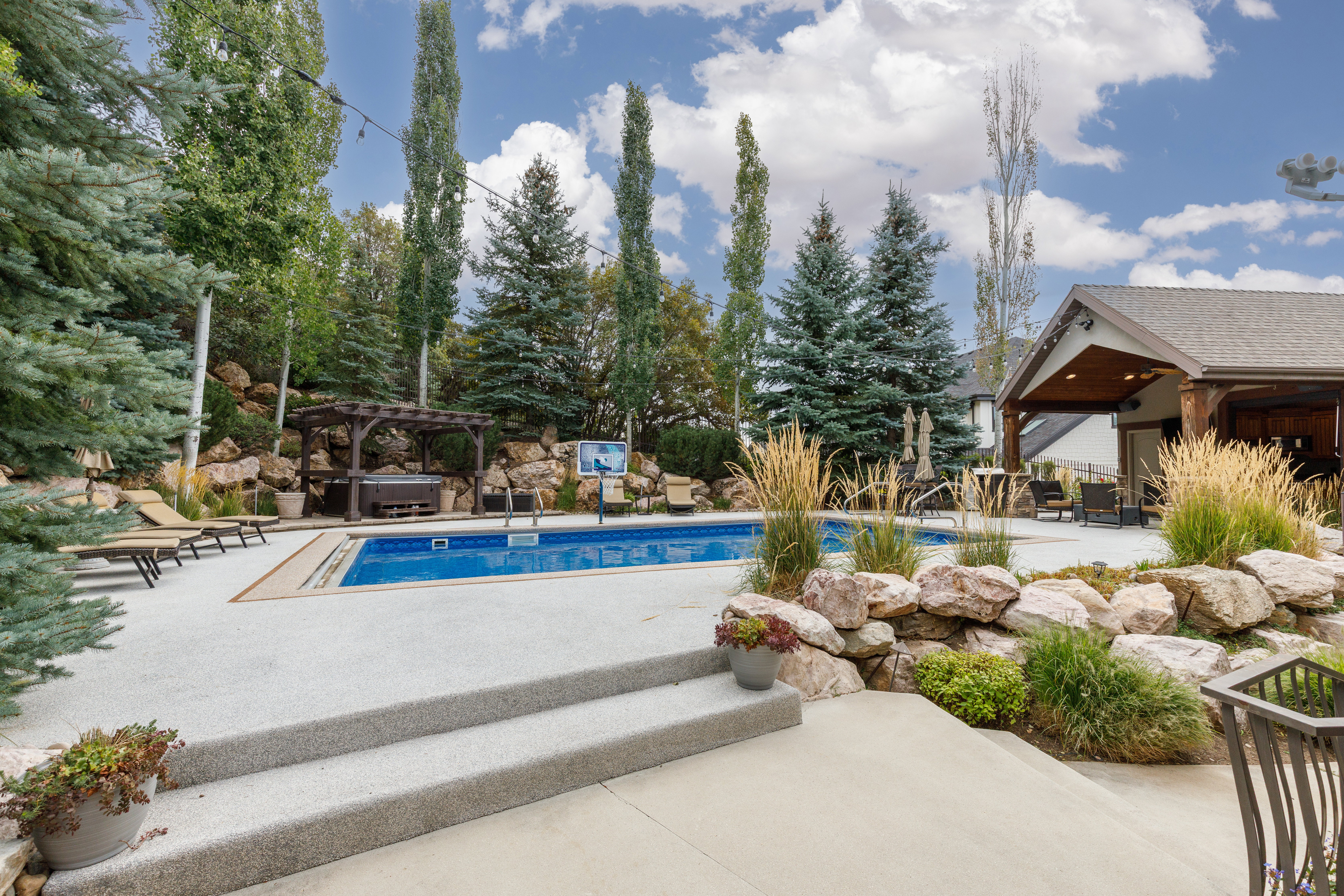 Outdoor entertaining space featuring a built-in swimming pool, a covered patio structure with exposed wood beams, a hot tub beneath a wooden pergola, and tiered landscaping with natural rock elements