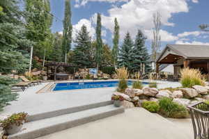 Outdoor entertaining space featuring a built-in swimming pool, a covered patio structure with exposed wood beams, a hot tub beneath a wooden pergola, and tiered landscaping with natural rock elements
