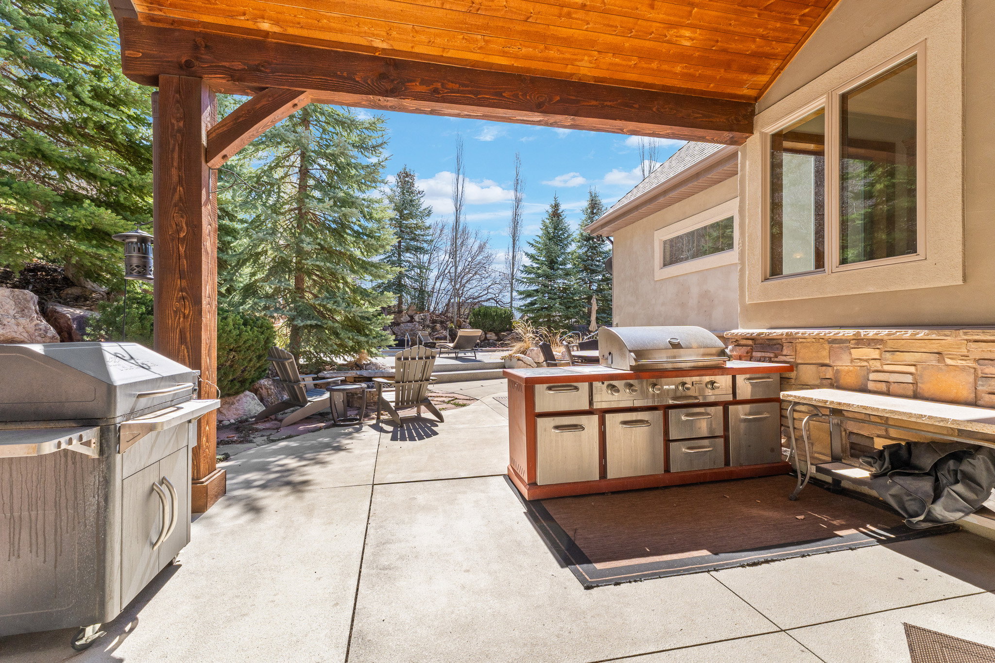 Covered patio featuring a built-in outdoor kitchen with stainless steel appliances and wood cabinetry, stone veneer accents, concrete flooring, and substantial wood beams