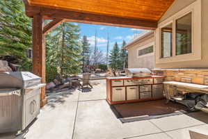 Covered patio featuring a built-in outdoor kitchen with stainless steel appliances and wood cabinetry, stone veneer accents, concrete flooring, and substantial wood beams