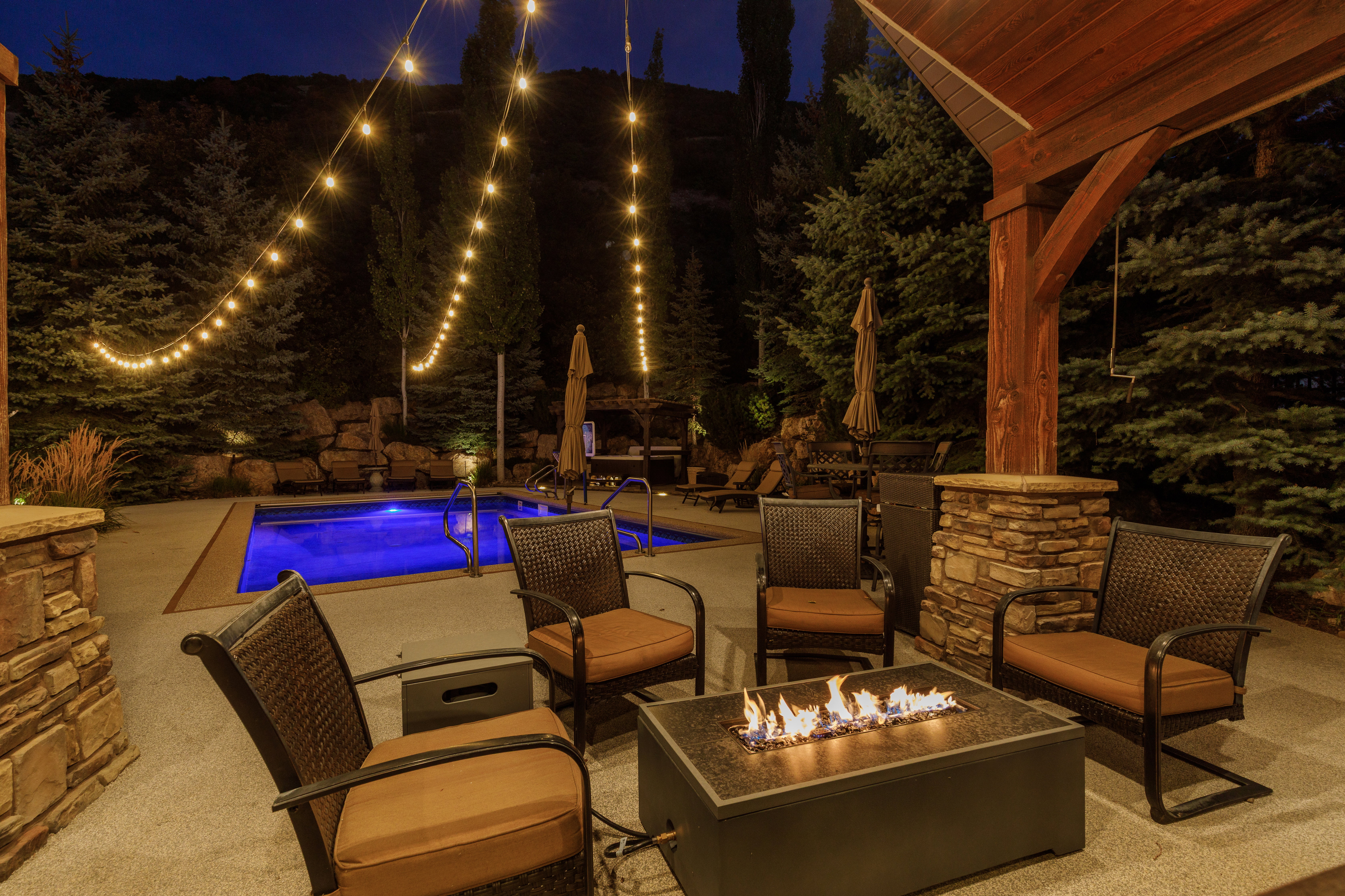 Outdoor entertainment area featuring a rectangular pool, stone-clad columns, a wood-beam pergola, and a built-in fire pit