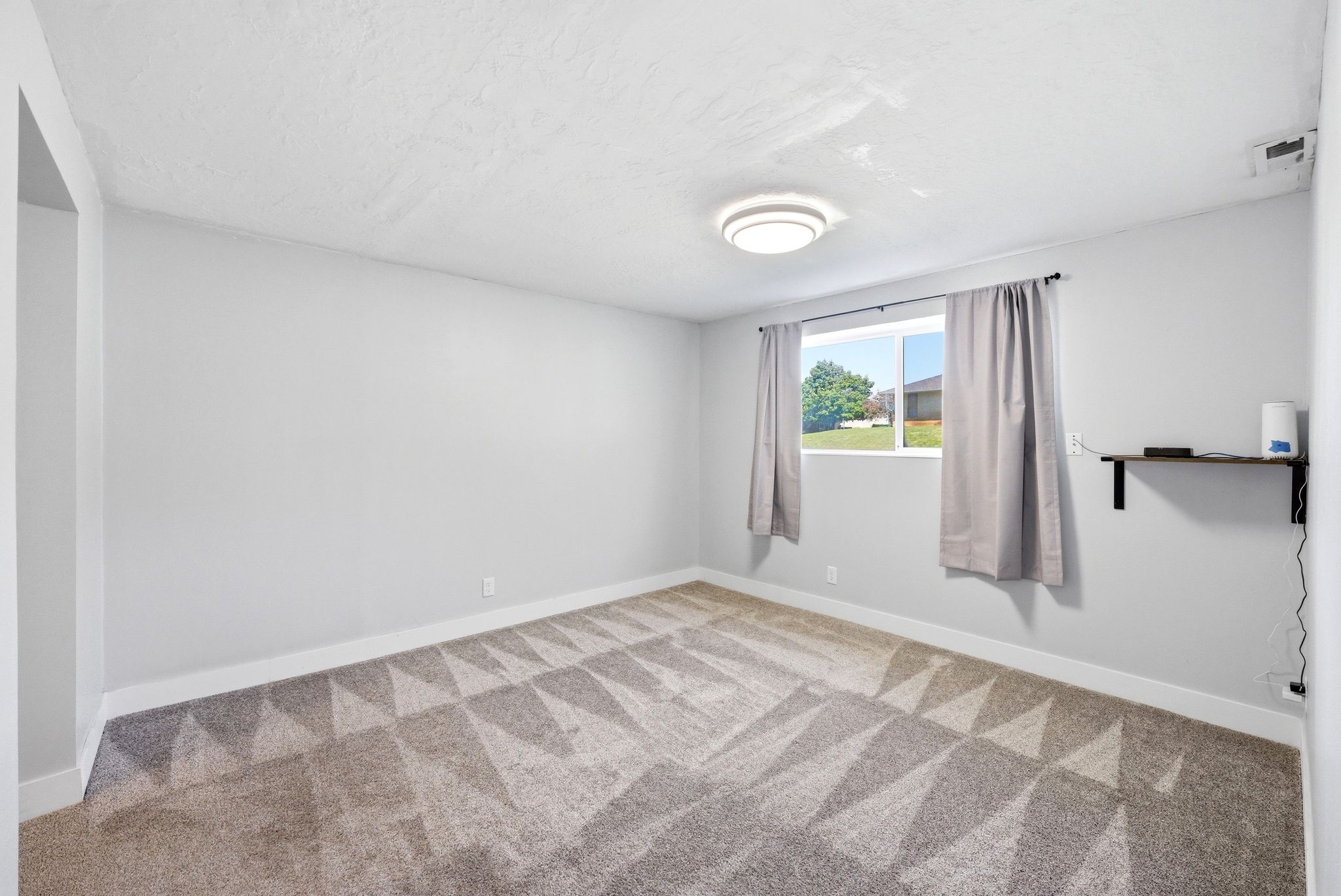 Spacious room featuring light gray walls, textured ceiling, geometric pattern carpeting, and a built-in wall shelf