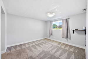 Spacious room featuring light gray walls, textured ceiling, geometric pattern carpeting, and a built-in wall shelf