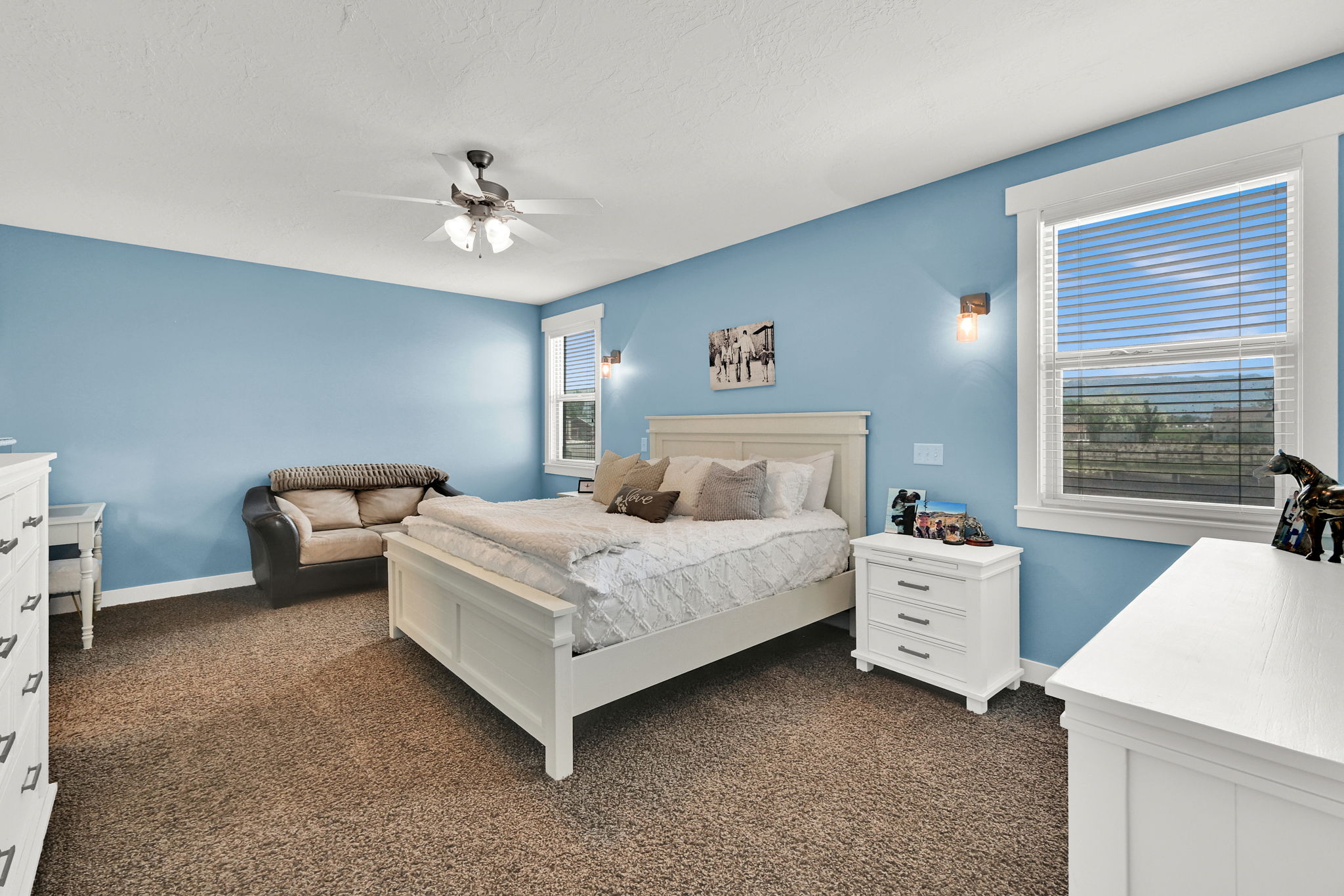 Carpeted room featuring a ceiling fan with integrated lighting, two windows with blinds, and wall-mounted sconce lighting