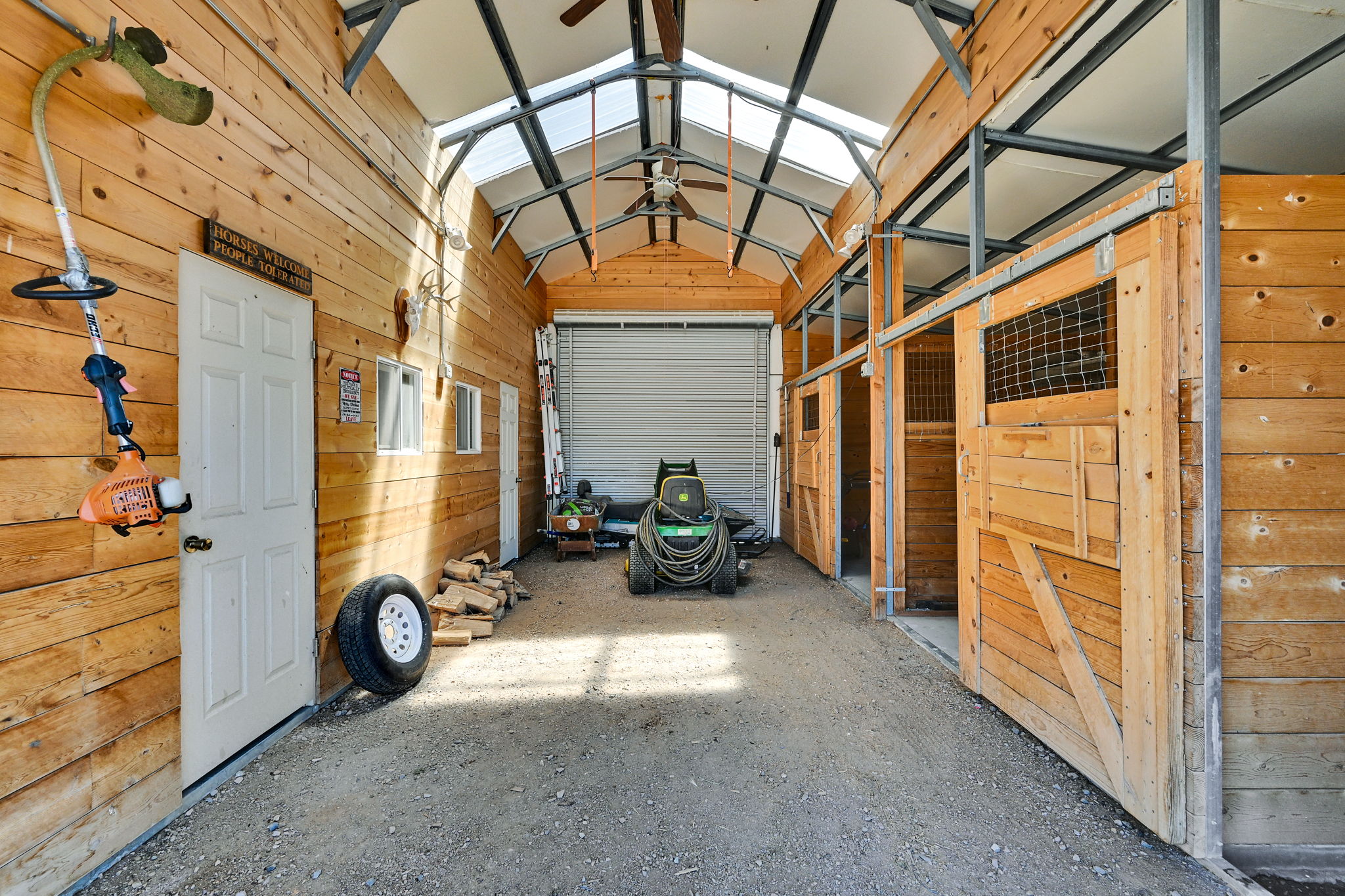 Spacious barn interior featuring natural wood plank walls, multiple sliding wooden stall doors with wire mesh inserts, exposed metal structural beams, a translucent roof panel for natural light, and a large roll-up door