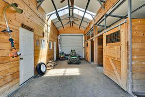 Spacious barn interior featuring natural wood plank walls, multiple sliding wooden stall doors with wire mesh inserts, exposed metal structural beams, a translucent roof panel for natural light, and a large roll-up door