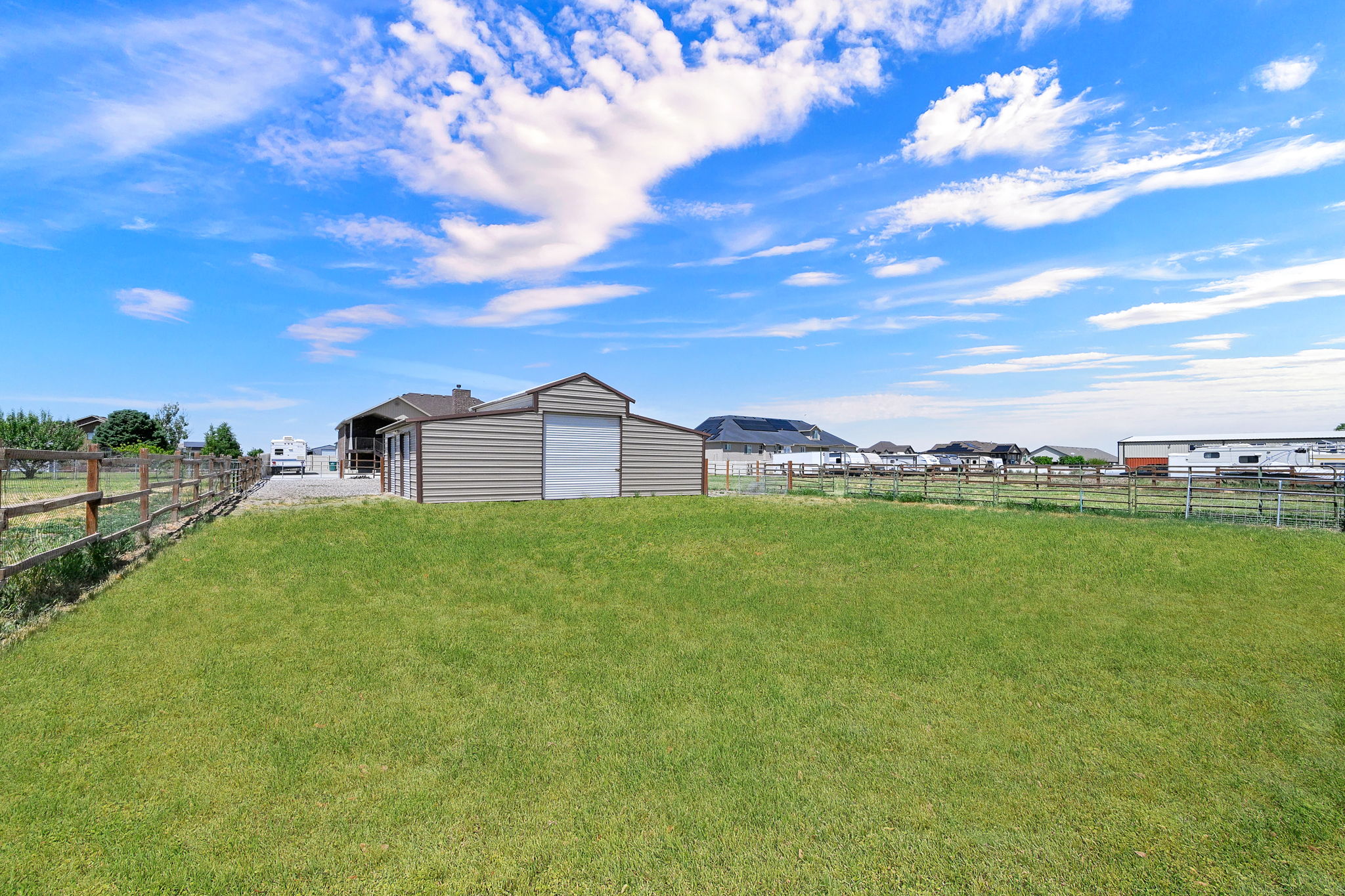 Expansive green lawn area featuring a split-rail fence, a metal outbuilding with an overhead door, and a covered patio structure