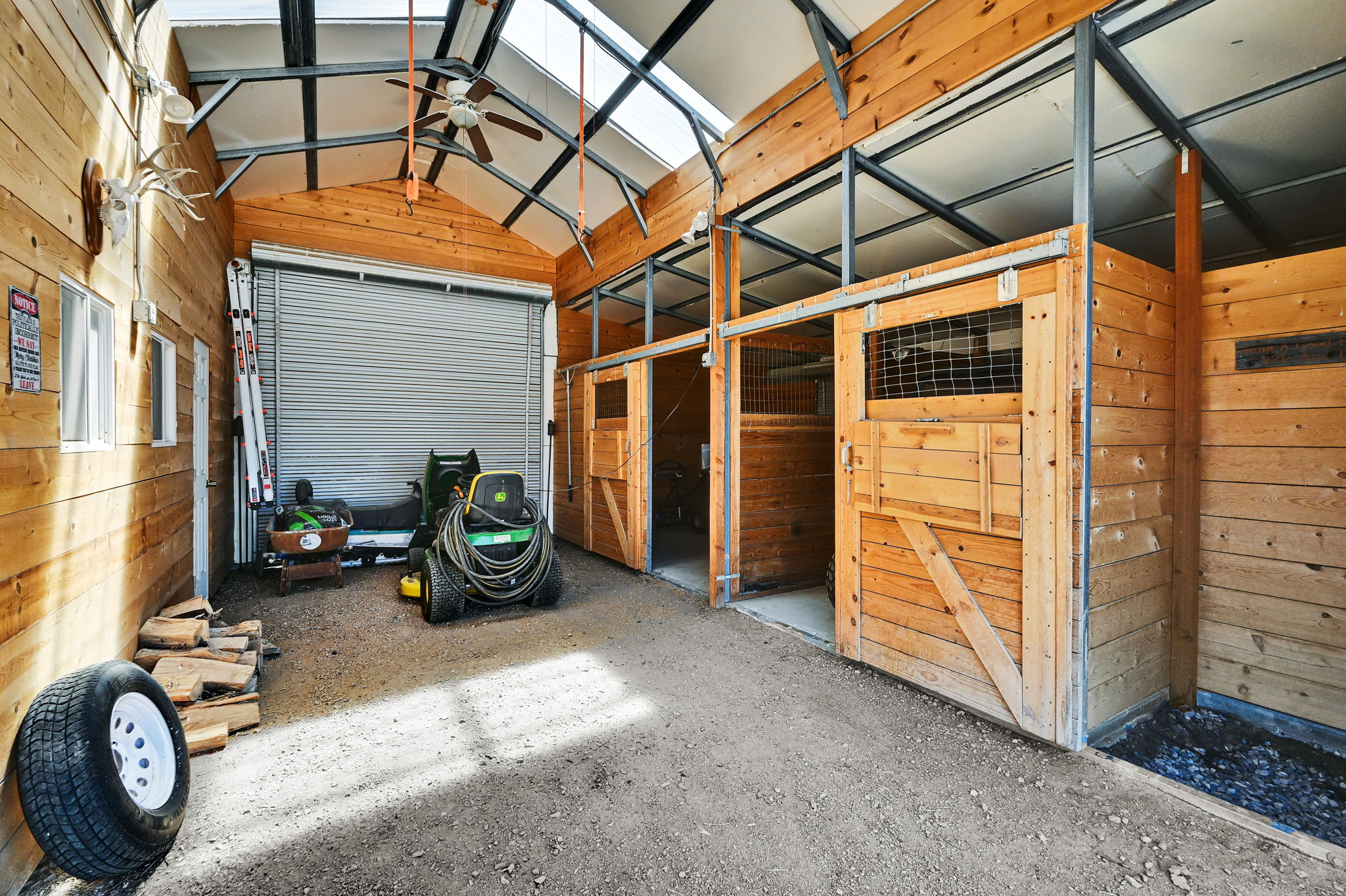 Interior structure featuring wood paneling, multiple barn-style stalls with sliding doors, a roll-up garage door, an exposed metal truss ceiling, and translucent roof panels
