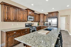 Kitchen featuring wood cabinetry, granite countertops, stainless steel appliances, a center island with a sink, and recessed lighting