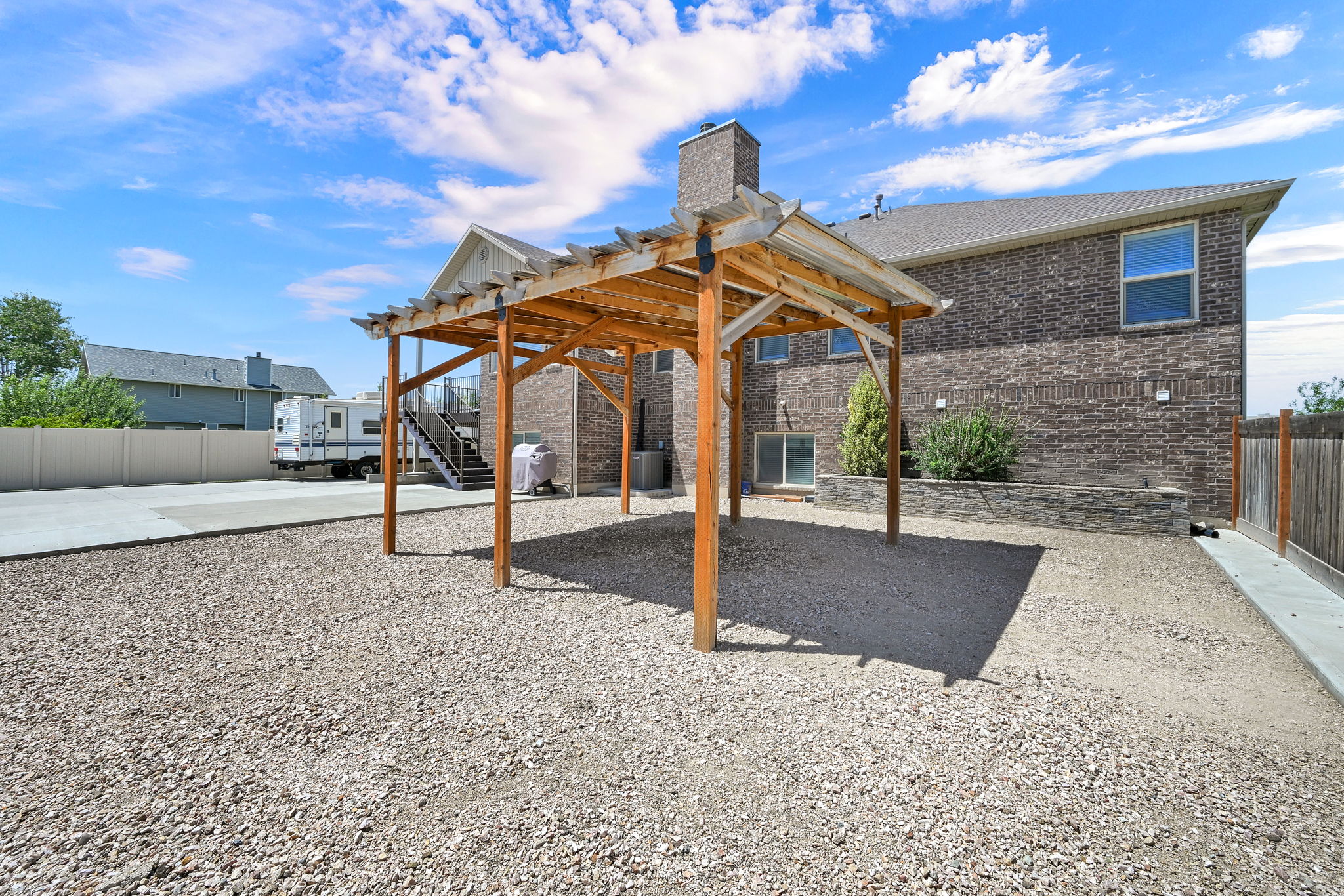 Spacious backyard featuring a wood pergola, gravel ground cover, brick exterior, stone retaining wall, and concrete pad
