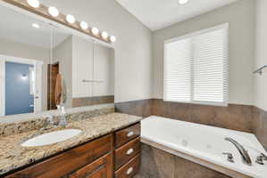 Bathroom featuring a built-in jetted tub with tile surround, wood cabinetry, granite countertop with integrated sink, and an expansive mirror with vanity lighting