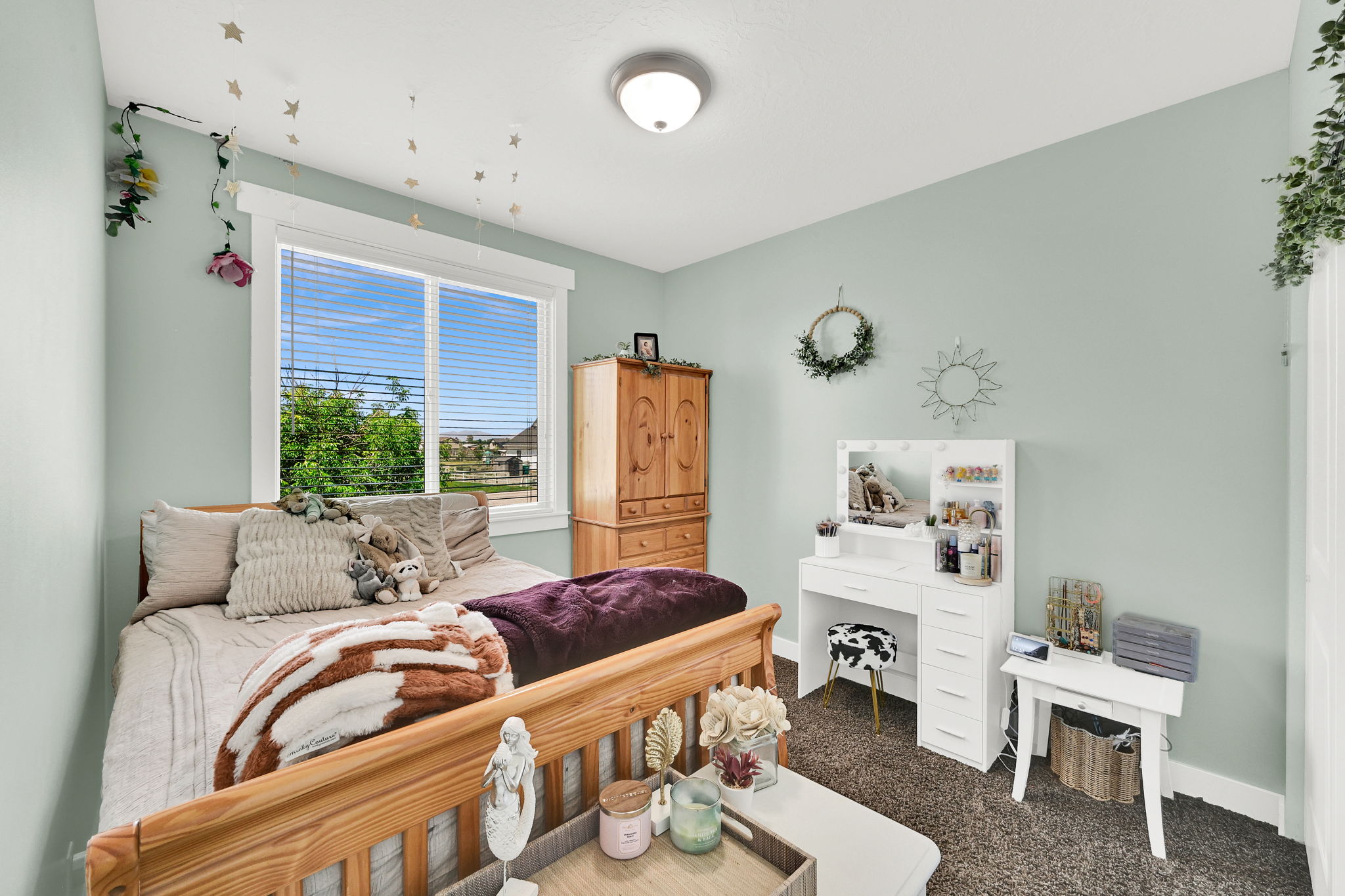 Single-pane window with white blinds, wood-finish bed frame, light green wall paint, overhead flush-mount light fixture, and dark carpet flooring