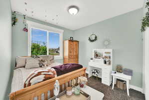 Single-pane window with white blinds, wood-finish bed frame, light green wall paint, overhead flush-mount light fixture, and dark carpet flooring