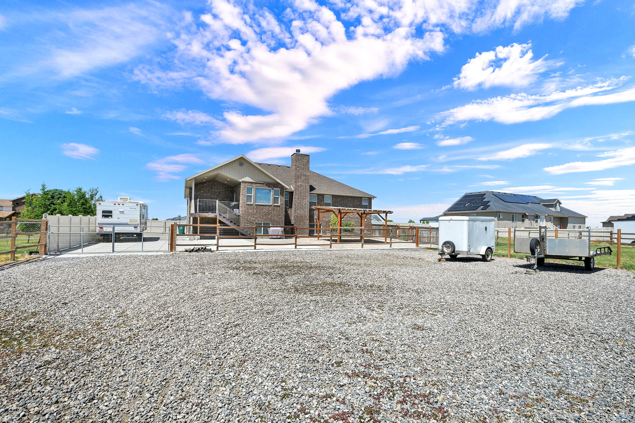 Expansive gravel yard area with a brick exterior structure featuring multiple windows, a second-story wooden deck with railing, and a ground-level patio with a wooden pergola