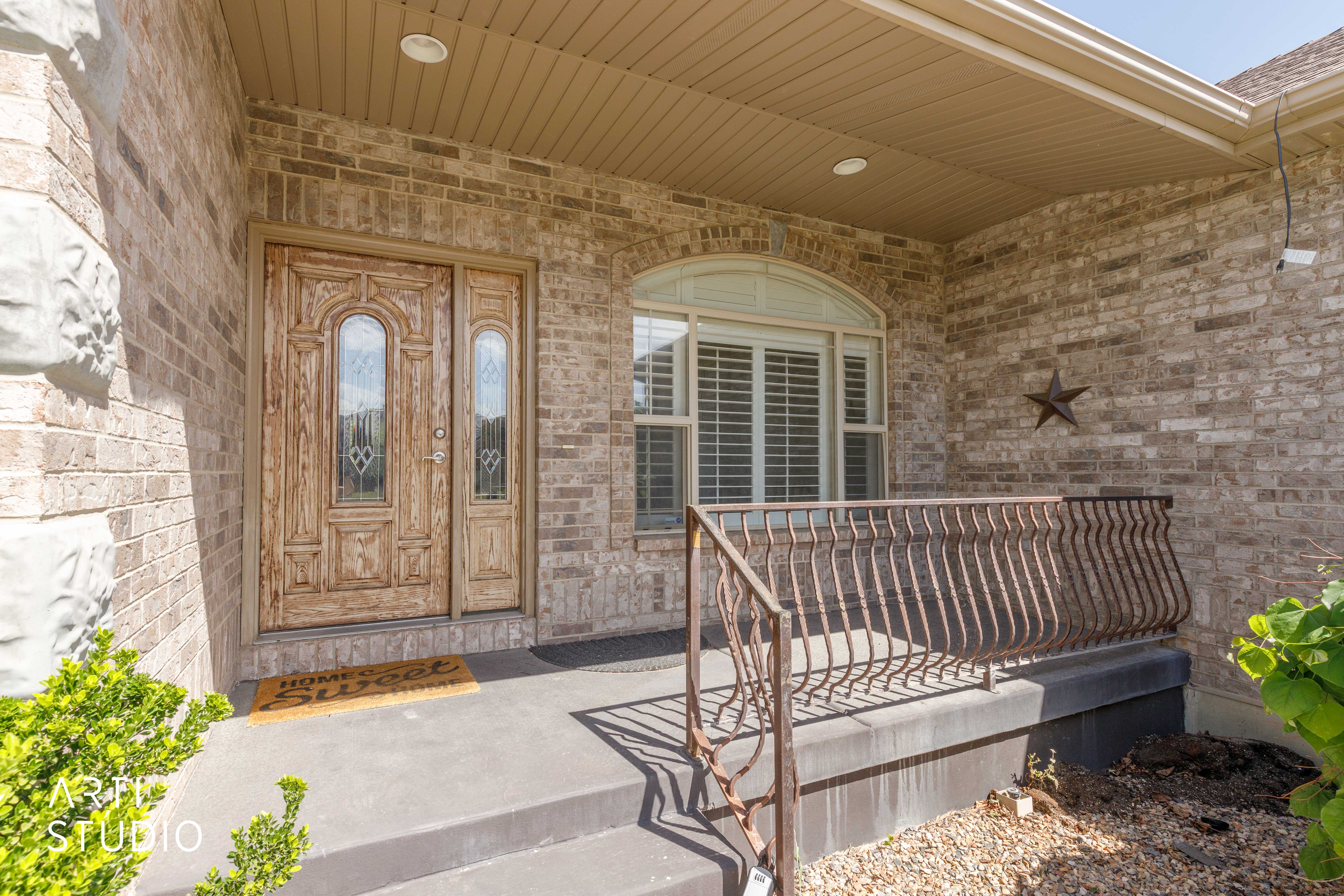 Brick exterior with a covered porch featuring a decorative wood-paneled door with glass inserts, an arched window with plantation shutters, and an ornate metal railing