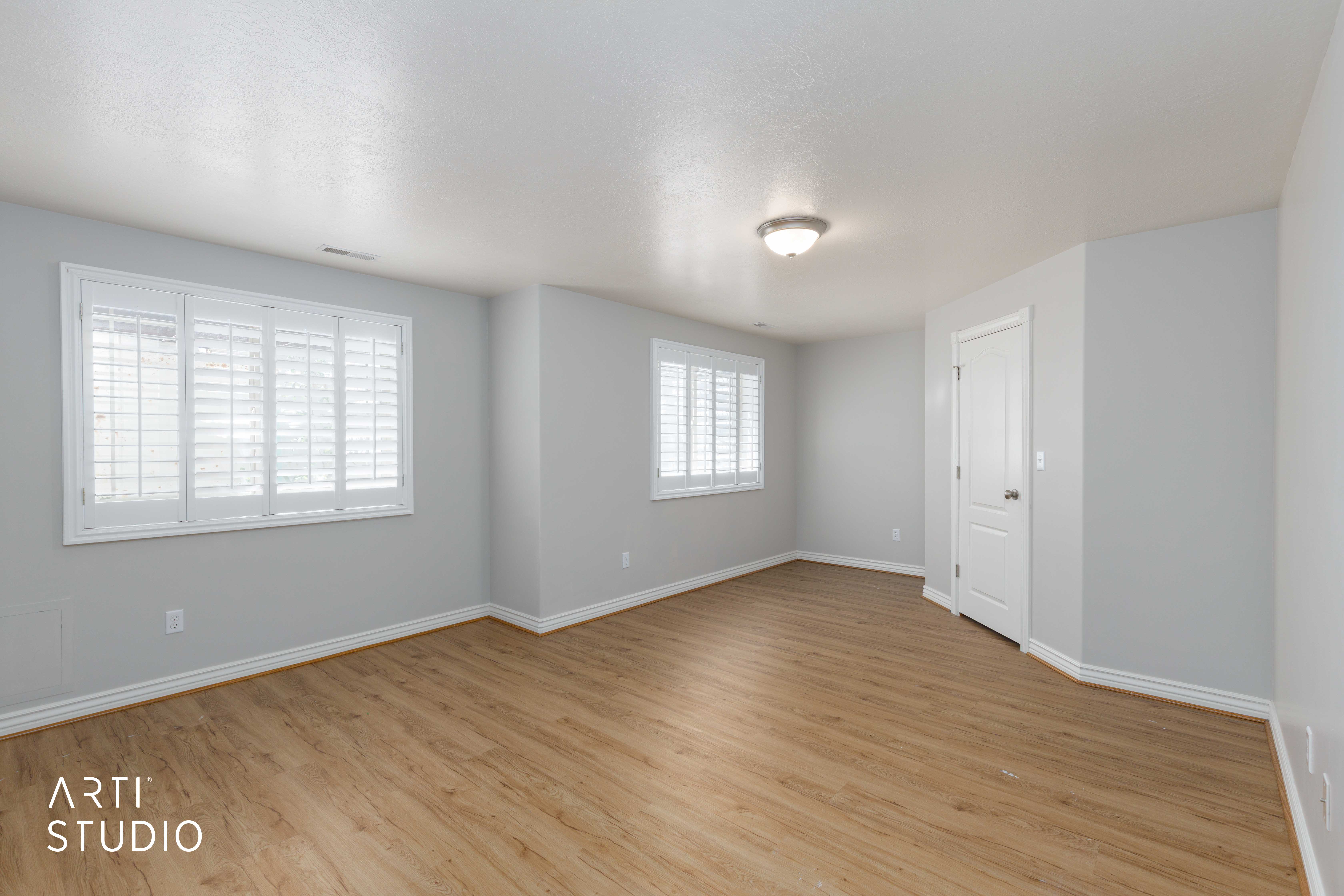 Spacious room featuring wood-finish flooring, light gray walls, white baseboards, and two windows with plantation shutters