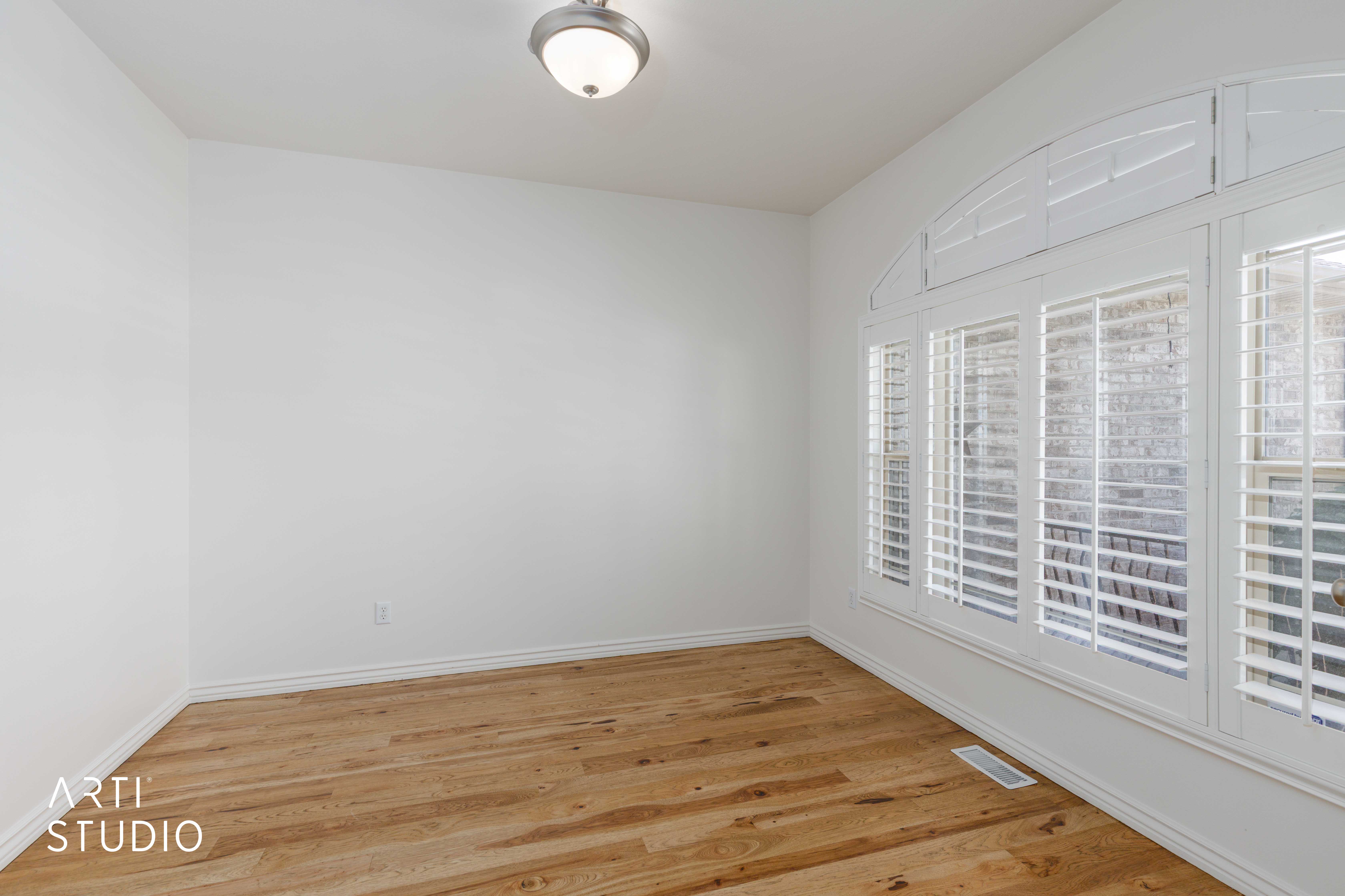Room featuring wood-finish flooring, white walls, and a multi-pane window with arched transom and plantation shutters