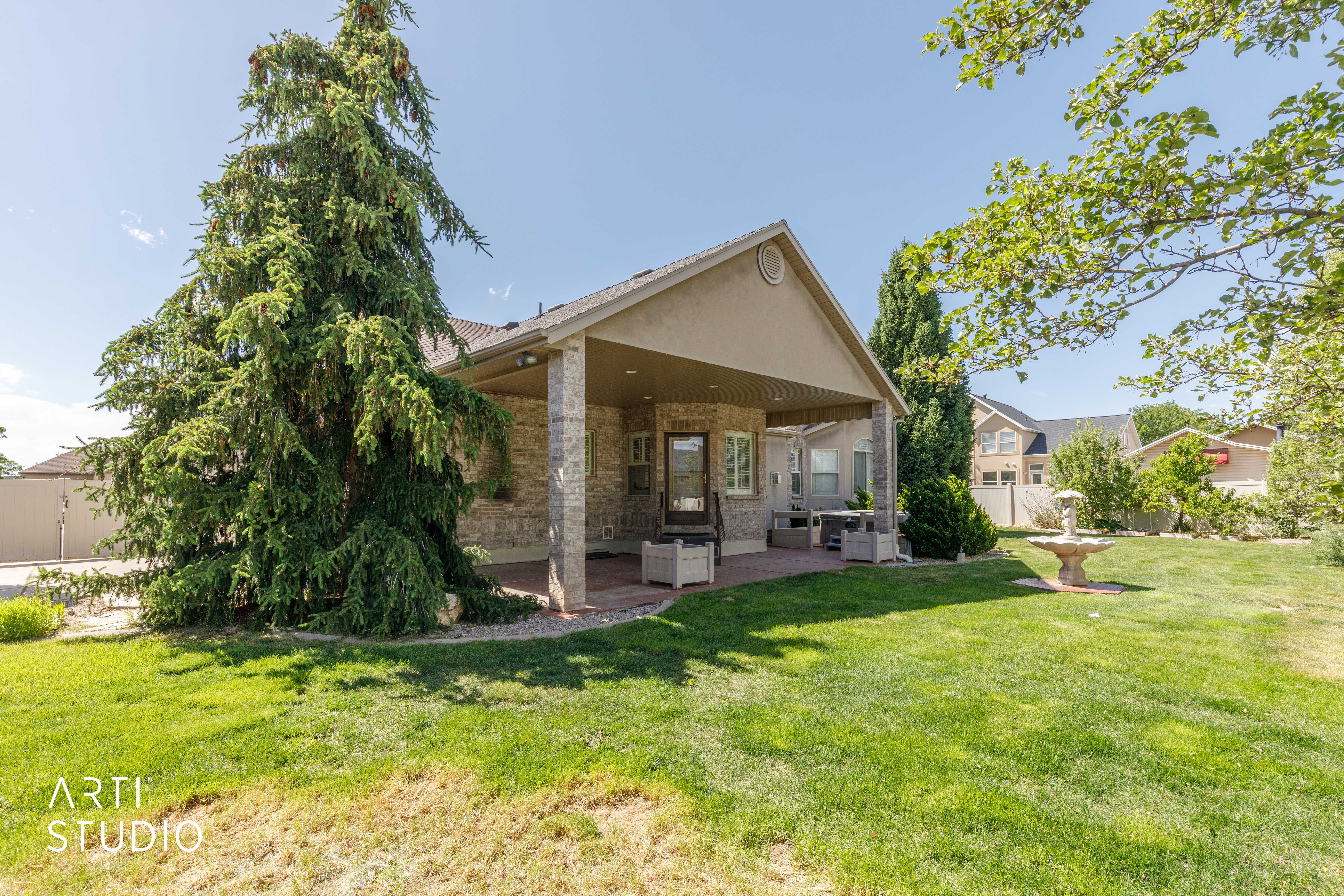 Expansive backyard featuring a covered patio with brick columns, a decorative fountain, and mature landscaping including a large evergreen tree