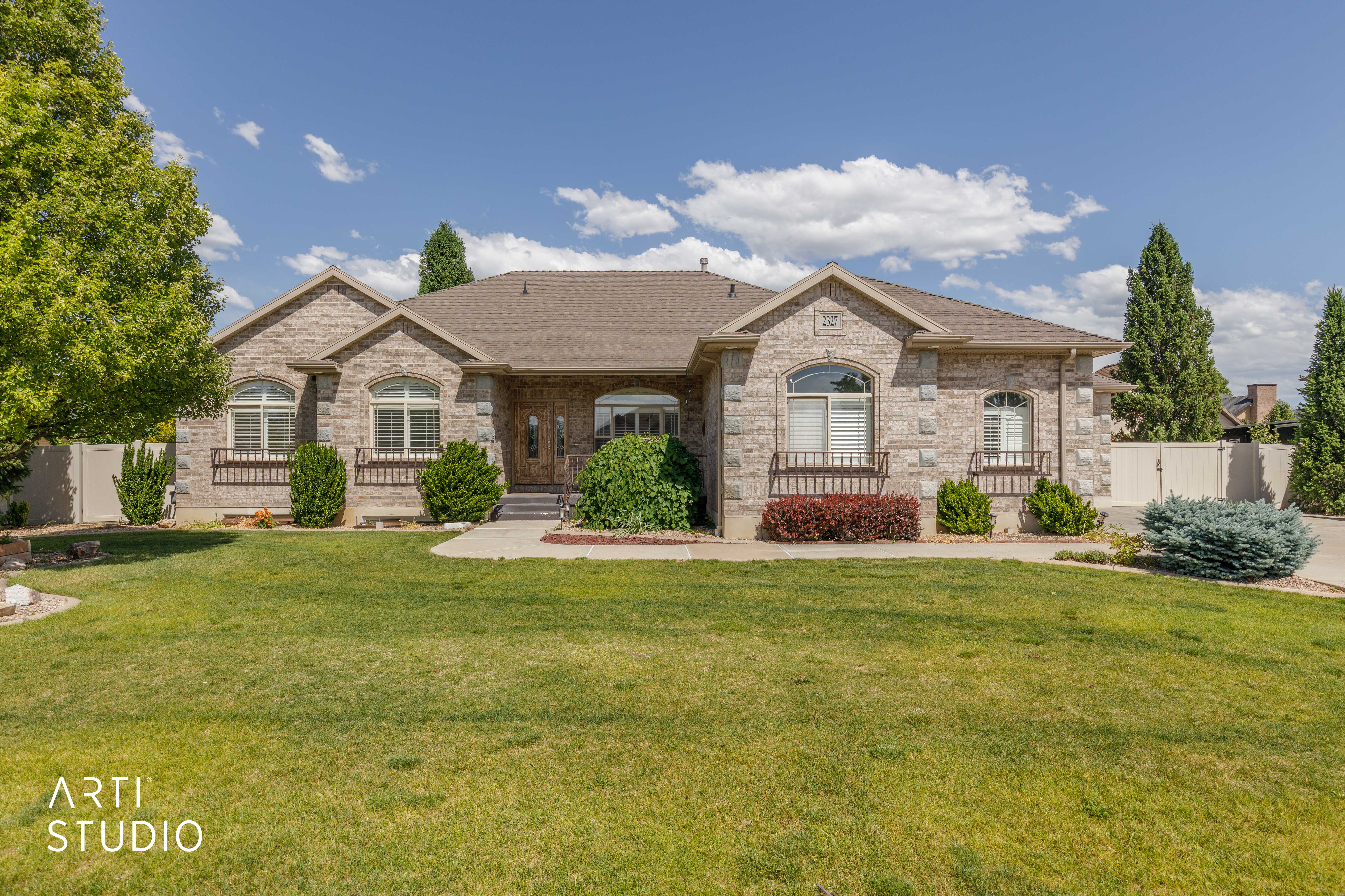 Stone veneer exterior with multiple arched windows, prominent gables, a dark shingle roof, and a maturely landscaped front yard with a paved driveway