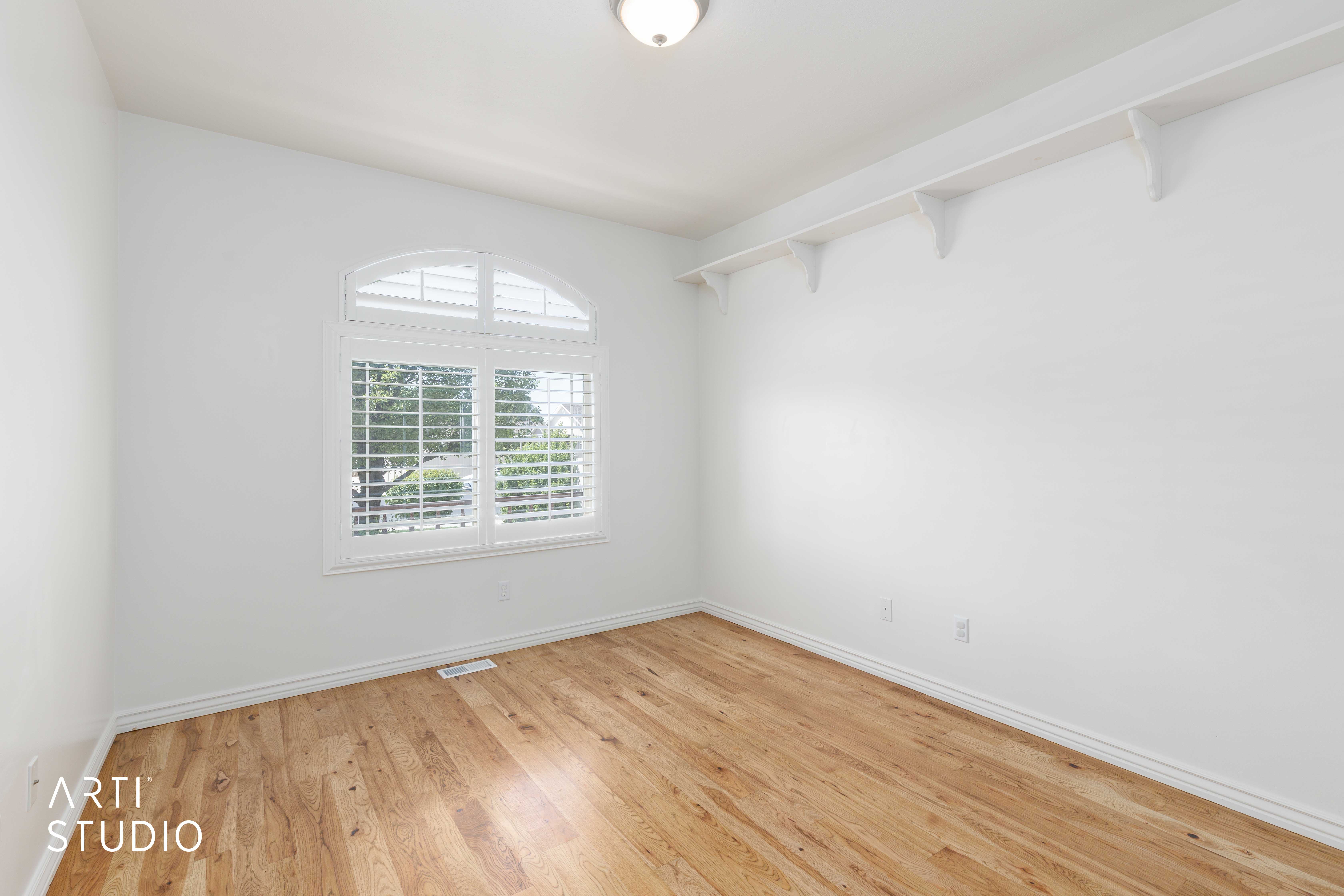 Bright interior space featuring wood-finish flooring, white walls, and an arched window with plantation shutters