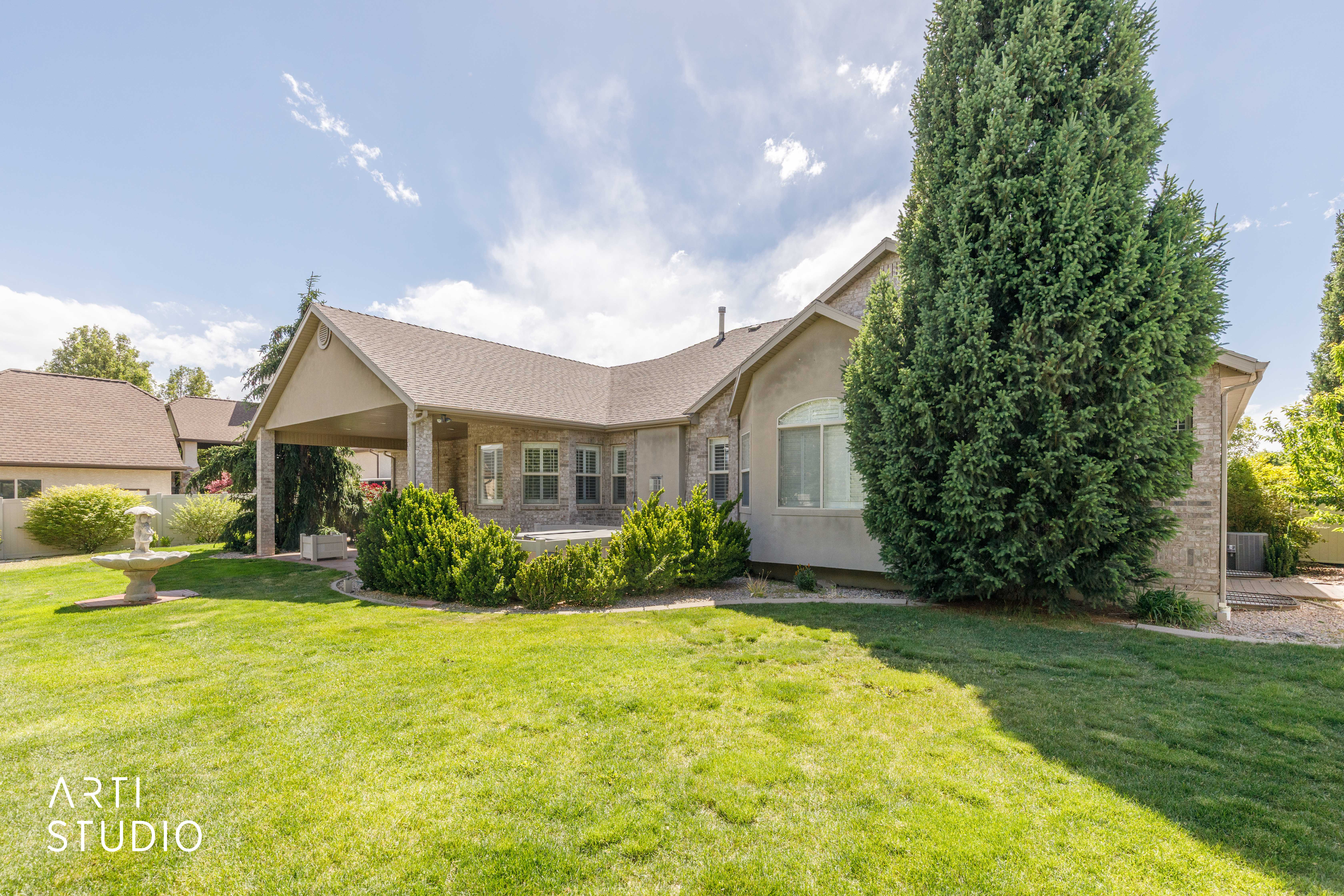 Expansive rear exterior featuring a covered patio, stone and stucco facade, multiple windows, and extensive landscaping with mature trees