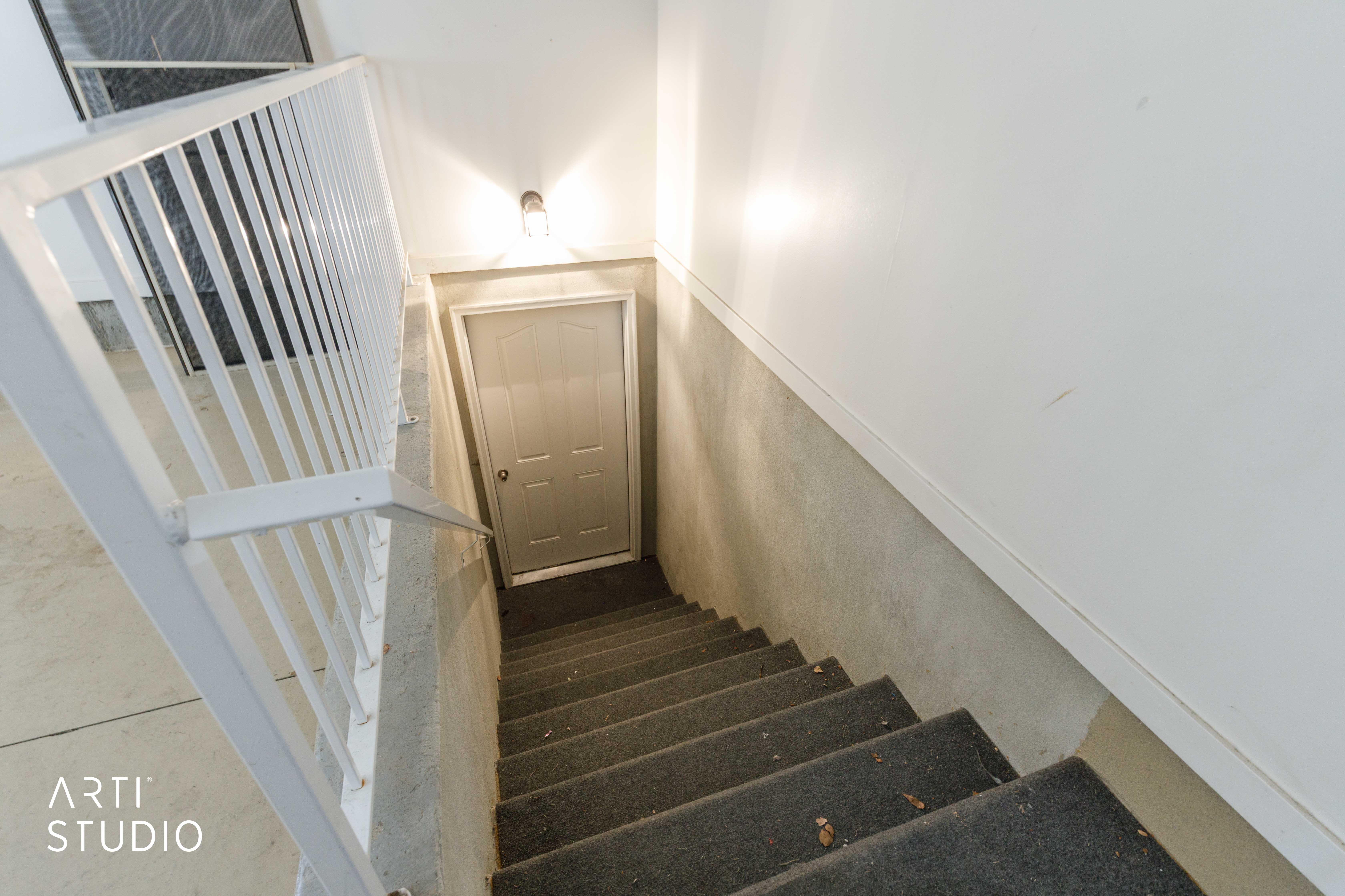 Carpeted staircase with white balusters and handrail, featuring a wall-mounted light fixture and a paneled door at the bottom