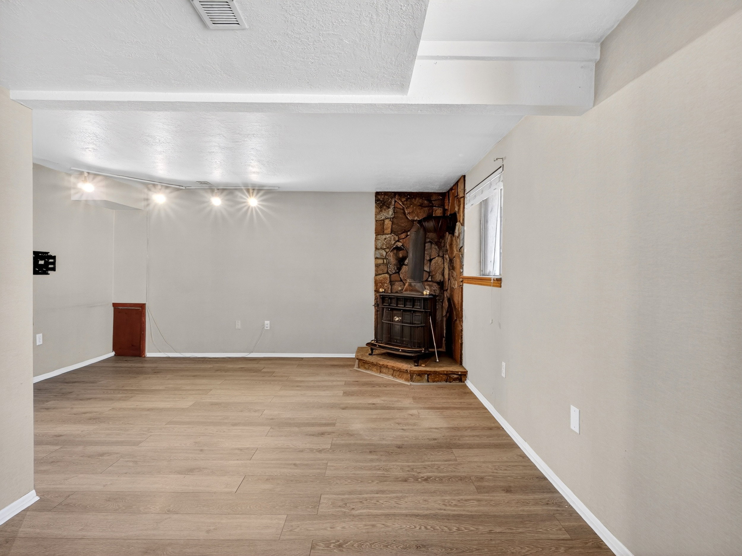Spacious room featuring wood-finish flooring, a stone-clad wood stove, track lighting, a window with wood trim, and painted walls