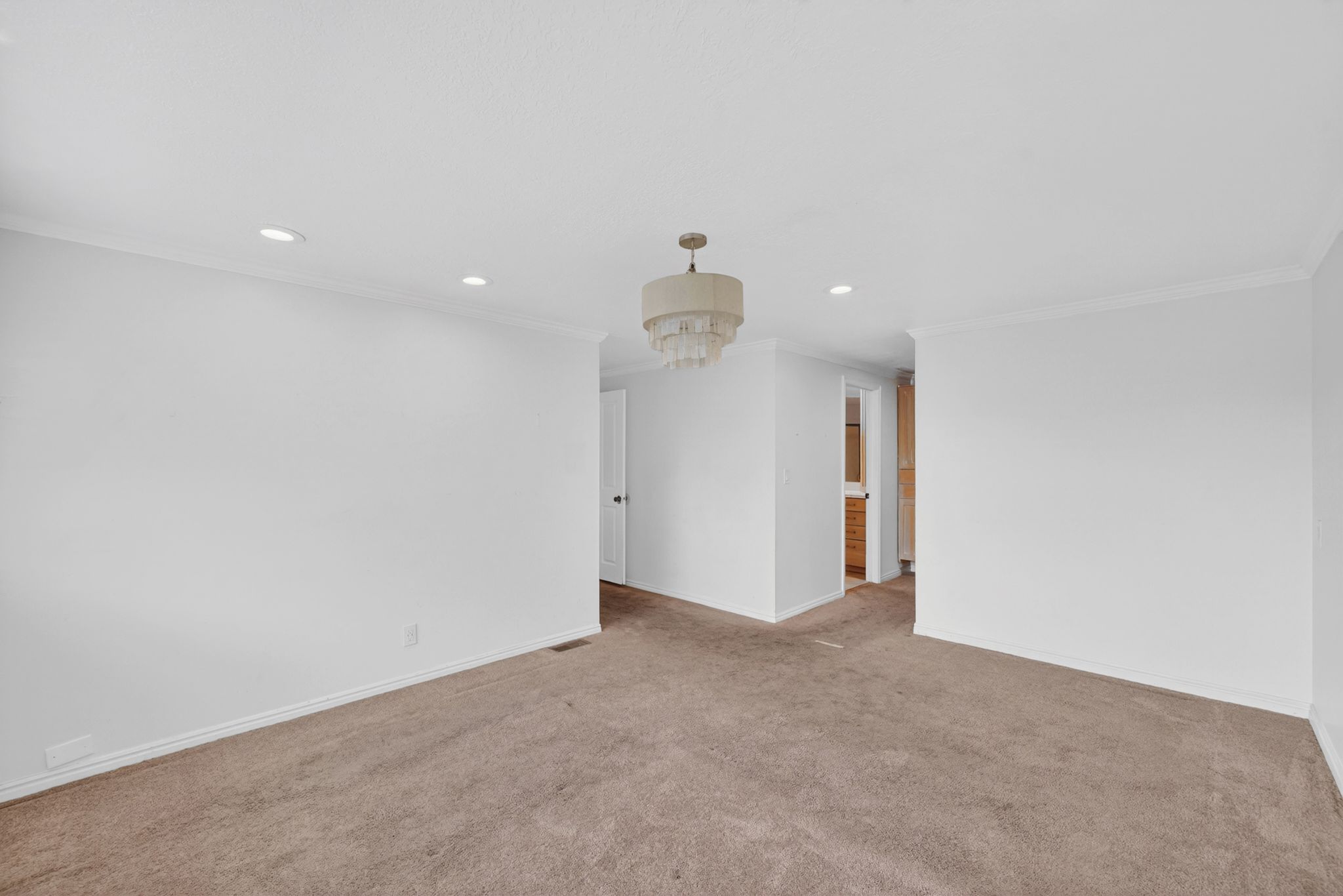 Spacious interior room featuring light brown carpeting, white walls with crown molding, recessed ceiling lighting, and a drum-style chandelier