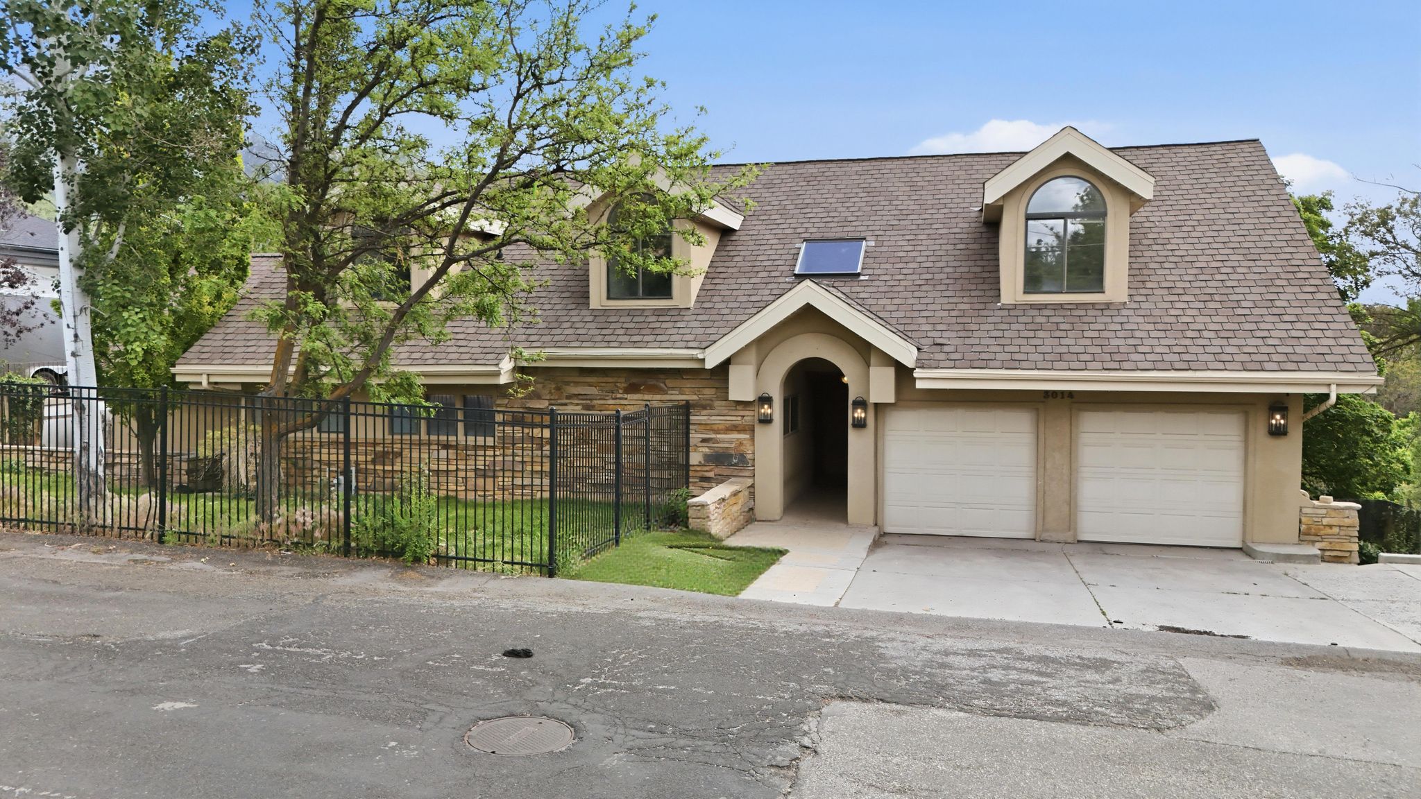 Stone veneer and stucco exterior with a two-car garage, dormer windows, and a black wrought iron fence