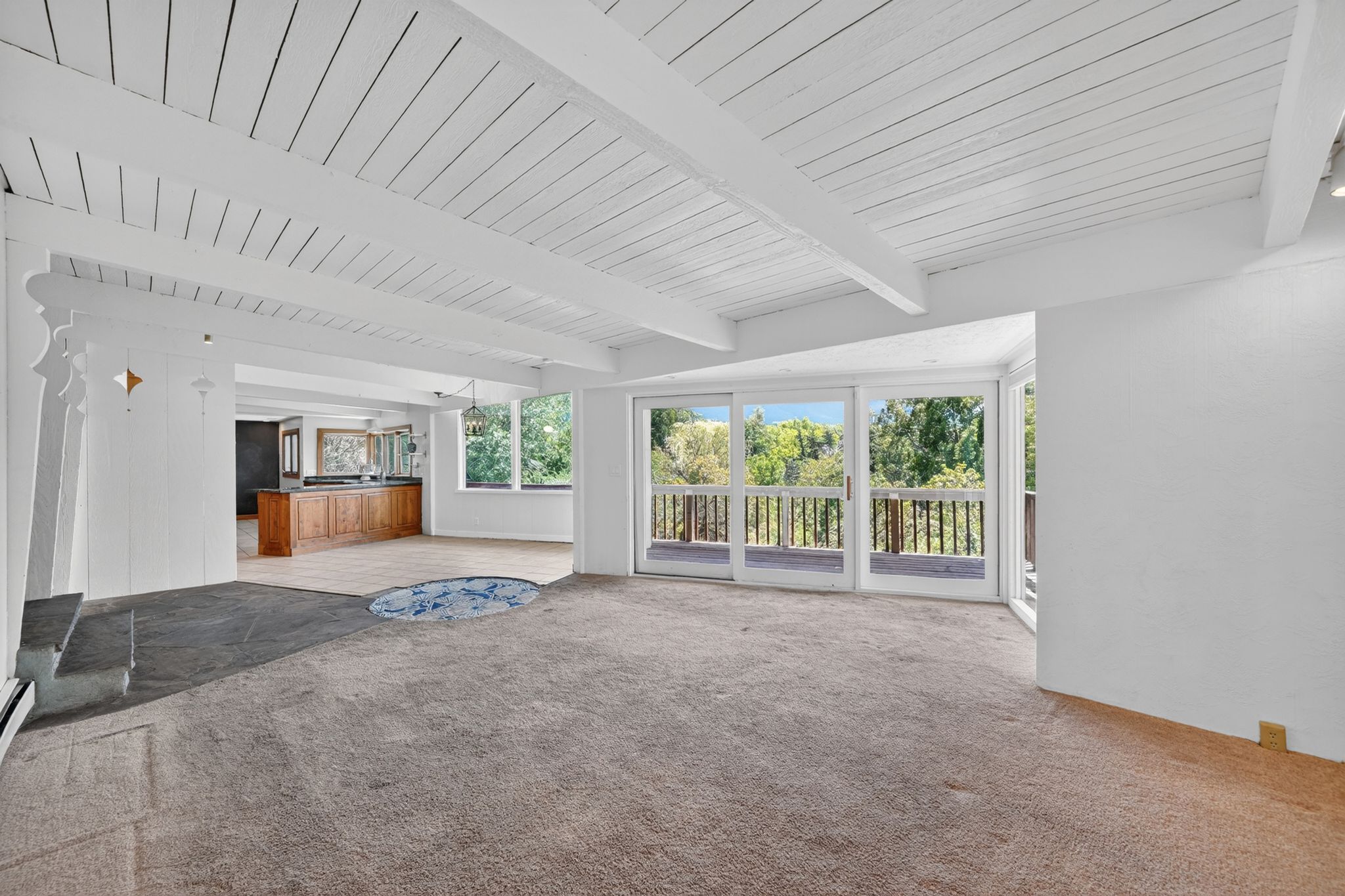 Open-concept living space featuring exposed white wood-beamed ceilings, light brown carpeting, and a stone hearth