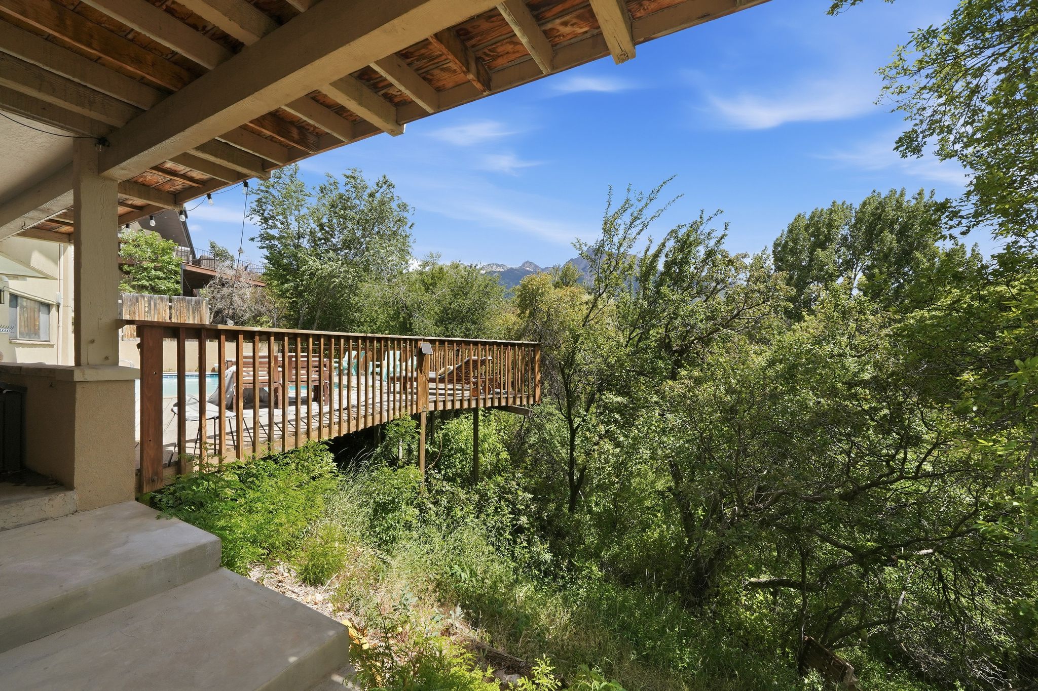 Covered outdoor patio area with concrete steps, wooden deck with railing, and lush green foliage