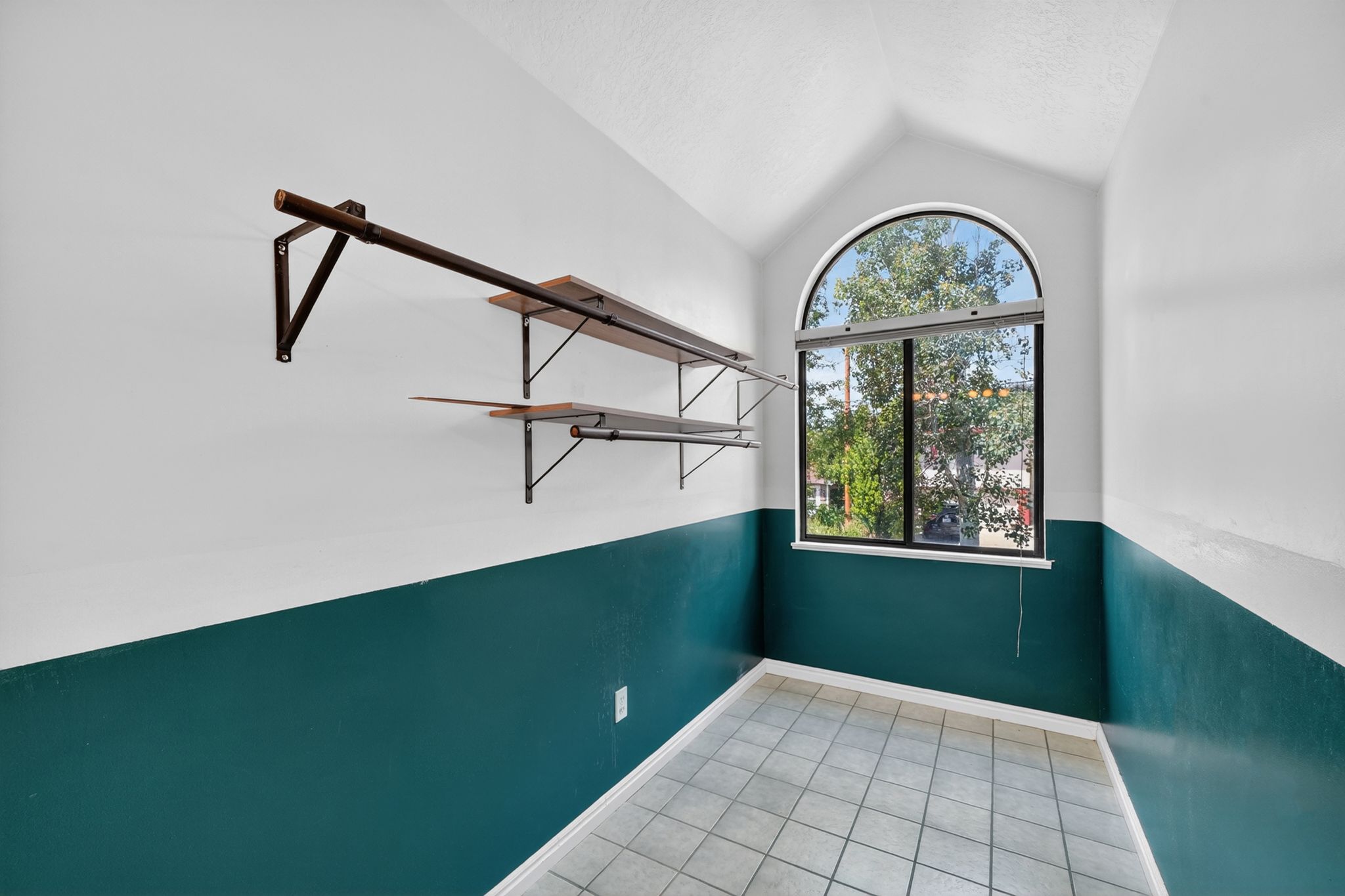 Room featuring a prominent arched window, vaulted ceiling, tile flooring, and wall-mounted shelving with a hanging rod