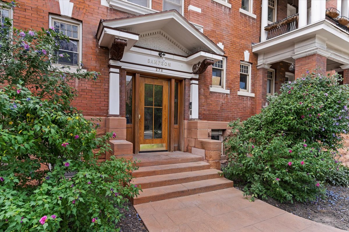Brick facade building entrance featuring a wood-panel glass-paned door, stone steps, a covered entryway with decorative brackets, and established shrubbery