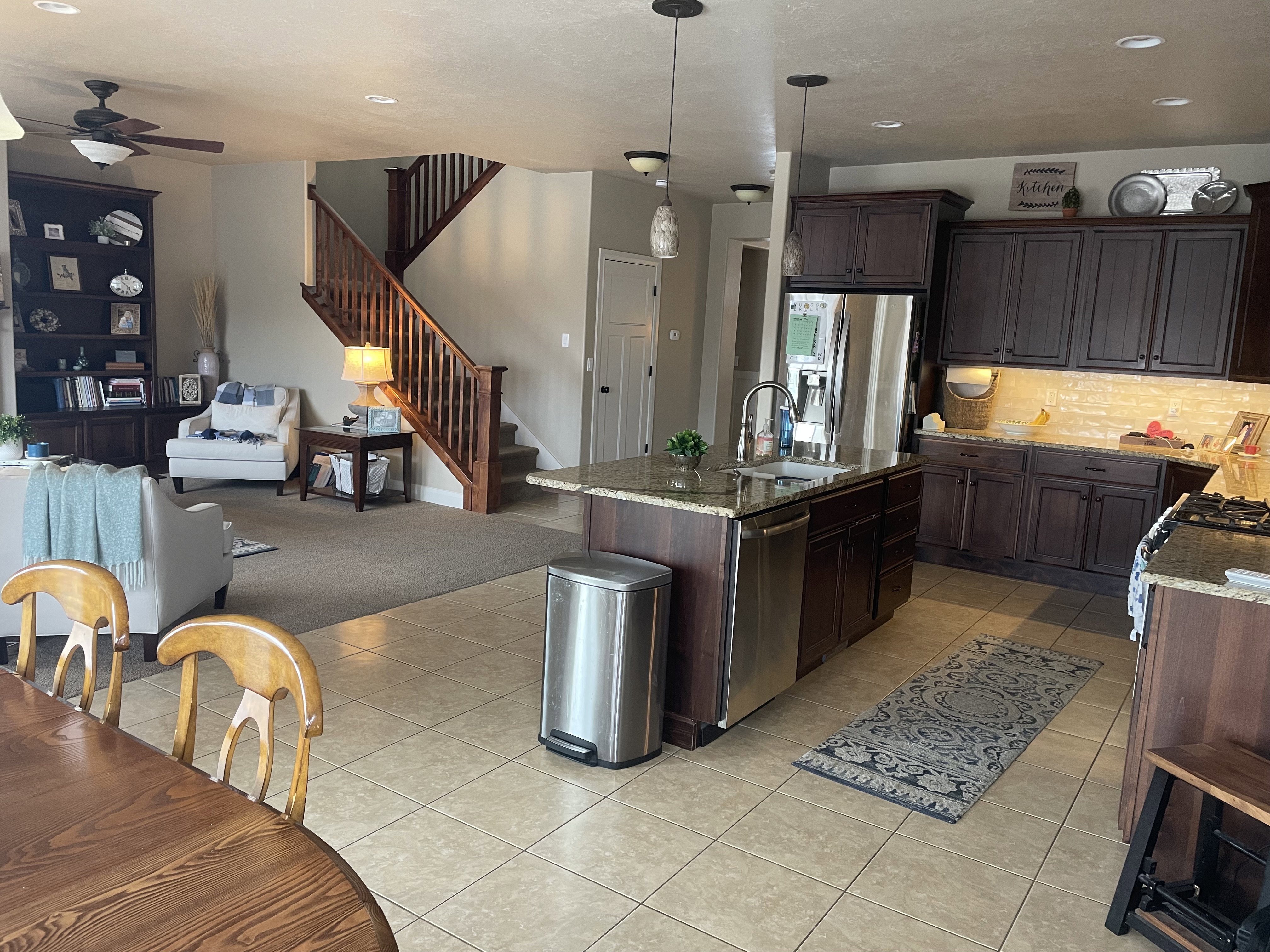 Open-concept kitchen and living area featuring tile flooring, a large kitchen island with integrated sink, dark wood cabinetry, and a carpeted living space with a visible staircase