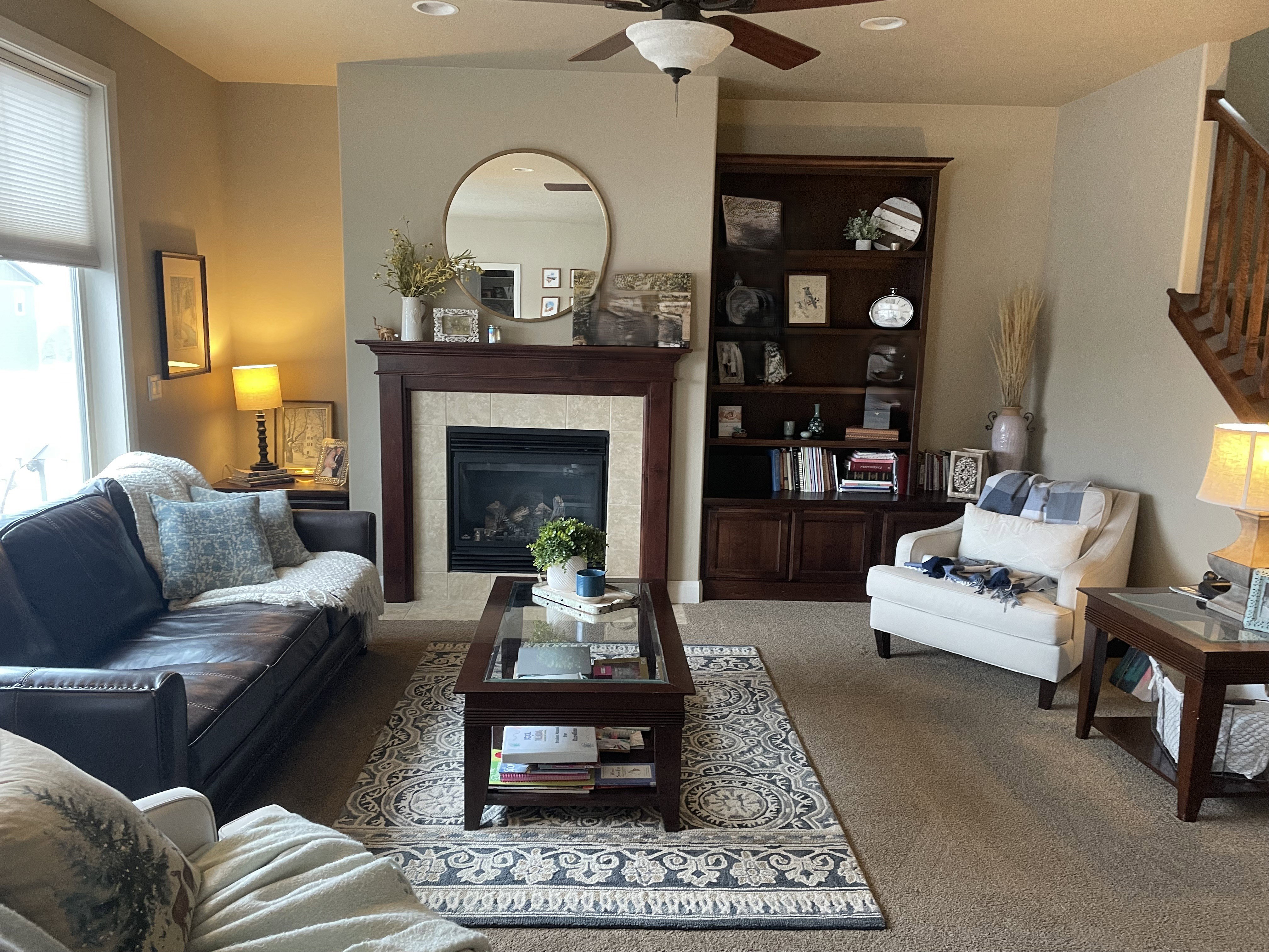 Living area featuring a gas fireplace with a tiled surround and wood mantel, built-in shelving, carpet flooring, a ceiling fan, and a prominent window with blinds