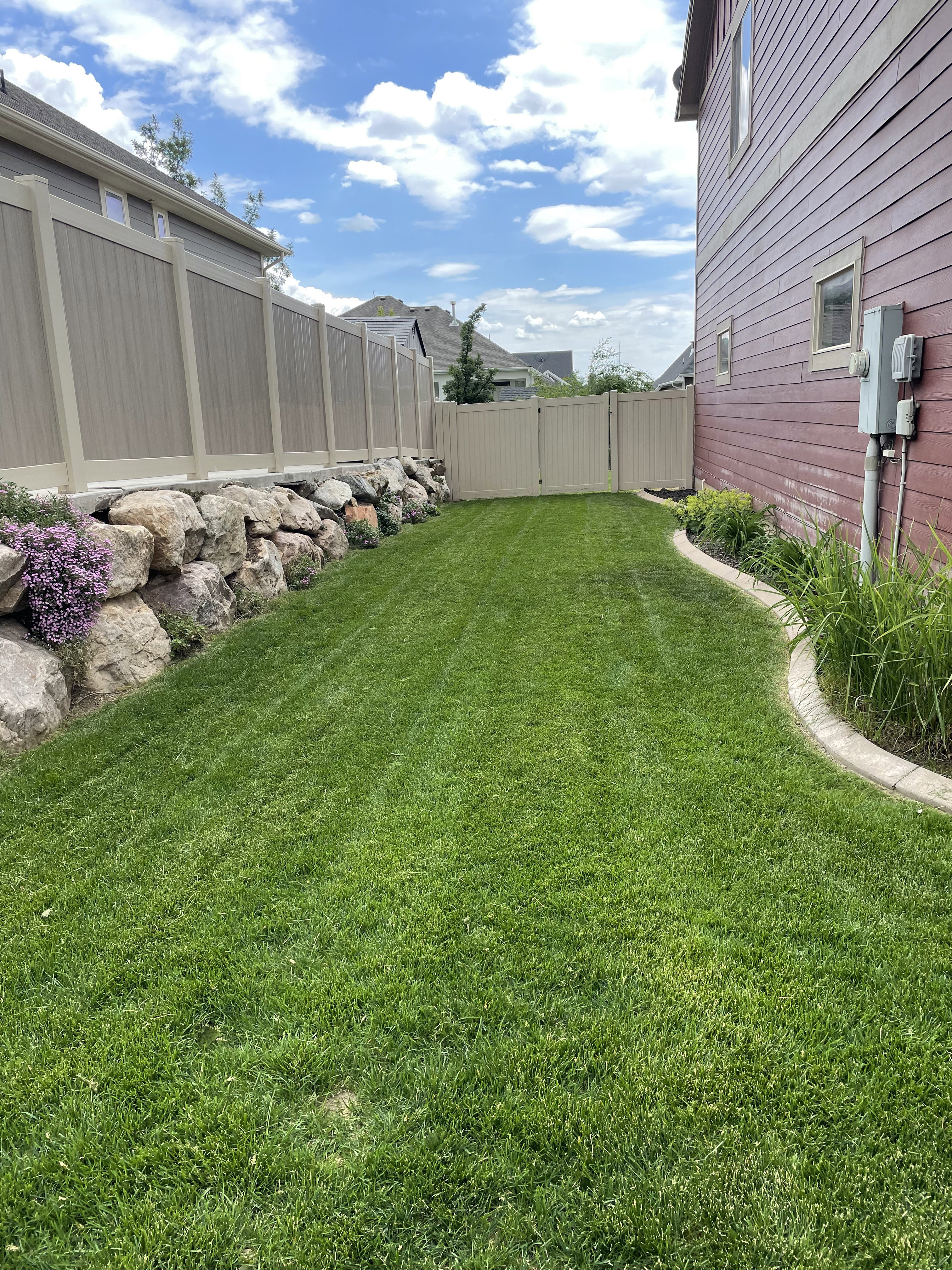 Expansive green lawn area featuring a decorative rock border with plantings, perimeter fencing, and a side access gate