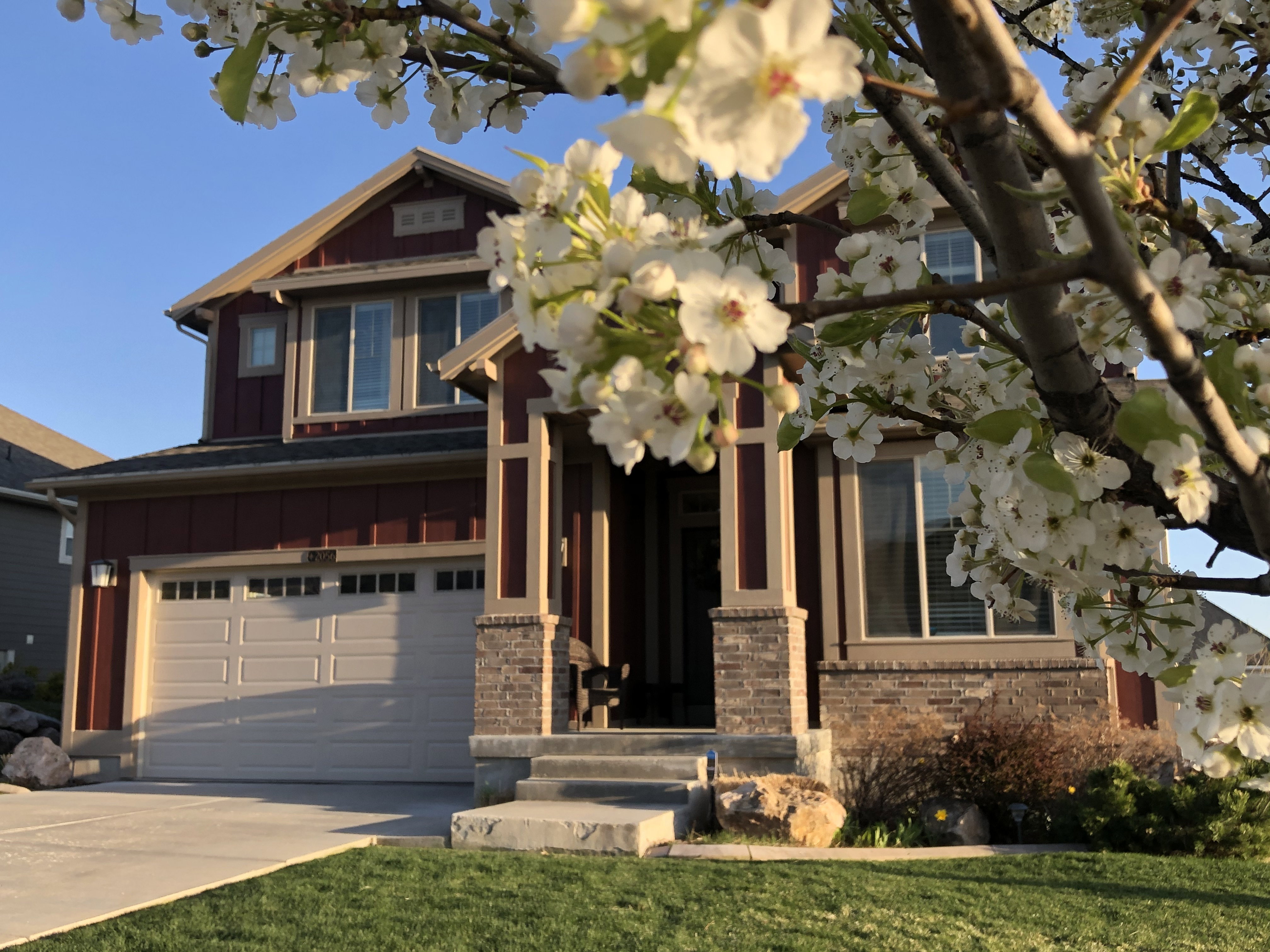 Two-story residence featuring a two-car garage with panel detailing, stone column accents, a covered front porch, and a landscaped front yard with mature plantings