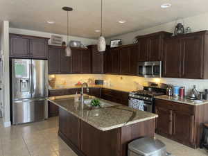 Kitchen featuring a central island with a sink, granite-style countertops, dark wood-finish cabinetry, stainless steel appliances, and recessed lighting