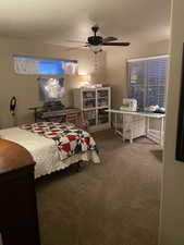 Carpeted room featuring a ceiling fan with integrated lighting, two windows with blinds, a built-in white shelving unit, and a dedicated workspace