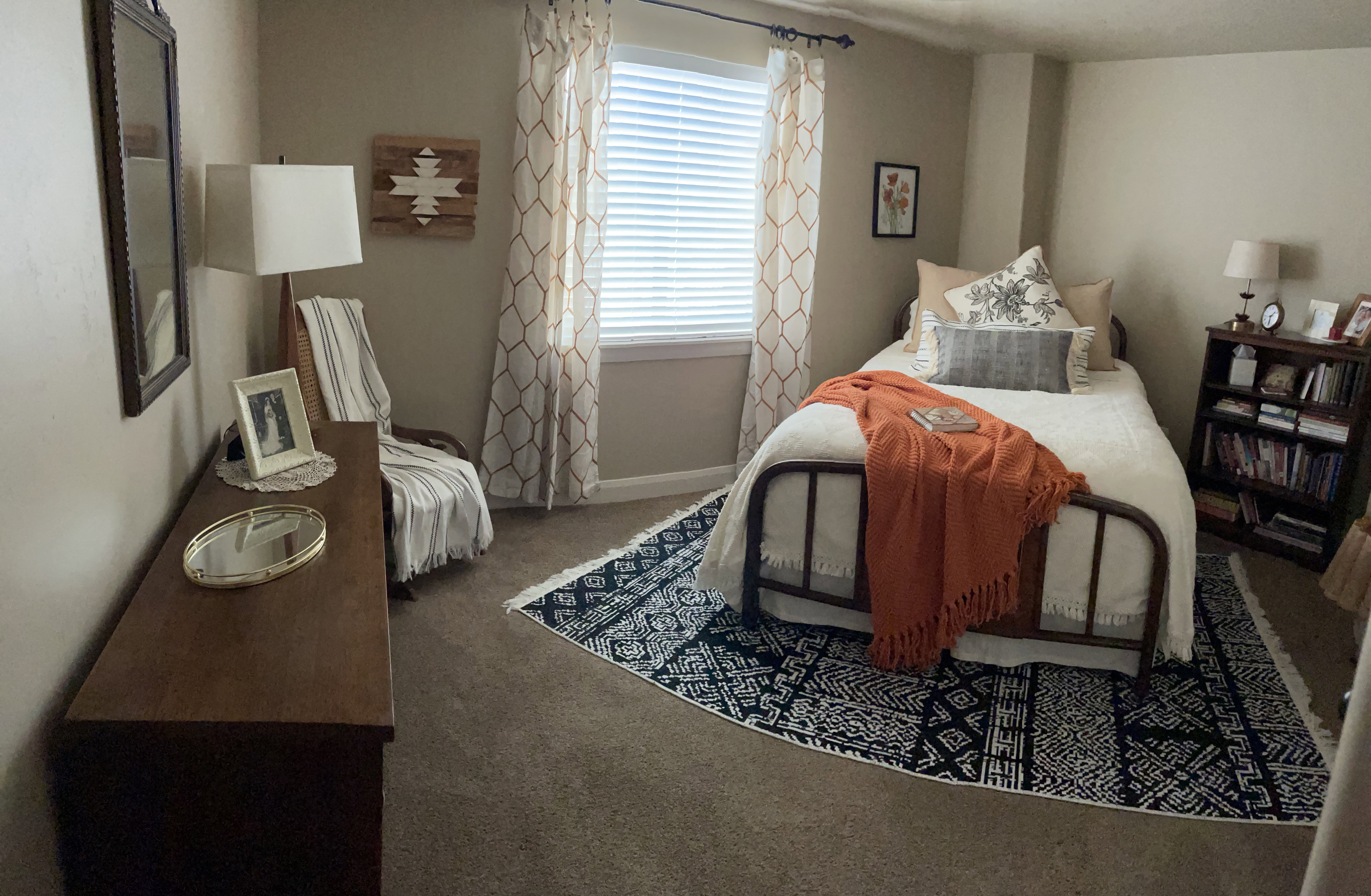 Carpeted room featuring a window with blinds and patterned drapes, a dark wood dresser, a dark wood bookshelf, and painted walls
