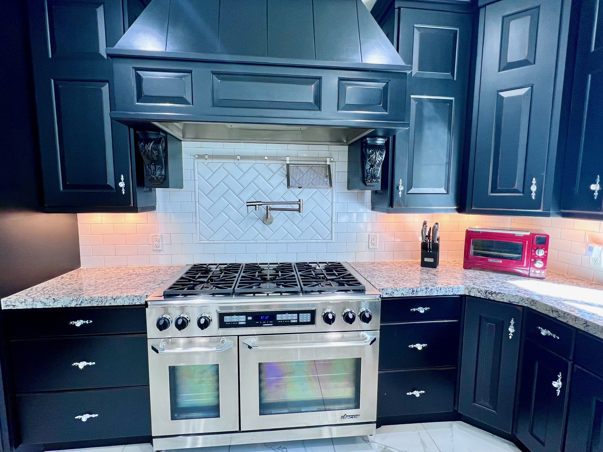 Gourmet kitchen featuring a professional-grade stainless steel range with double ovens, dark cabinetry with decorative hardware, light granite countertops, a subway tile backsplash, and a pot filler